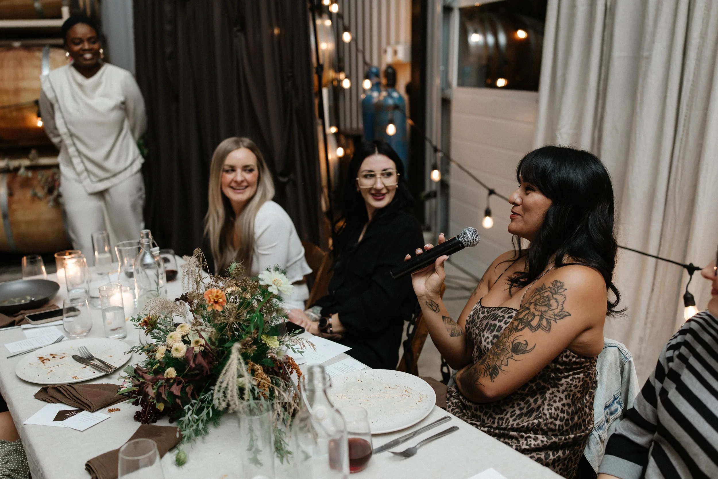 A woman with black hair and tattoos on her arm is speaking into a microphone at a decorated dinner table with three women listening attentively. The table has floral centerpieces, plates, glasses, and candles. The setting appears to be a cozy, warmly