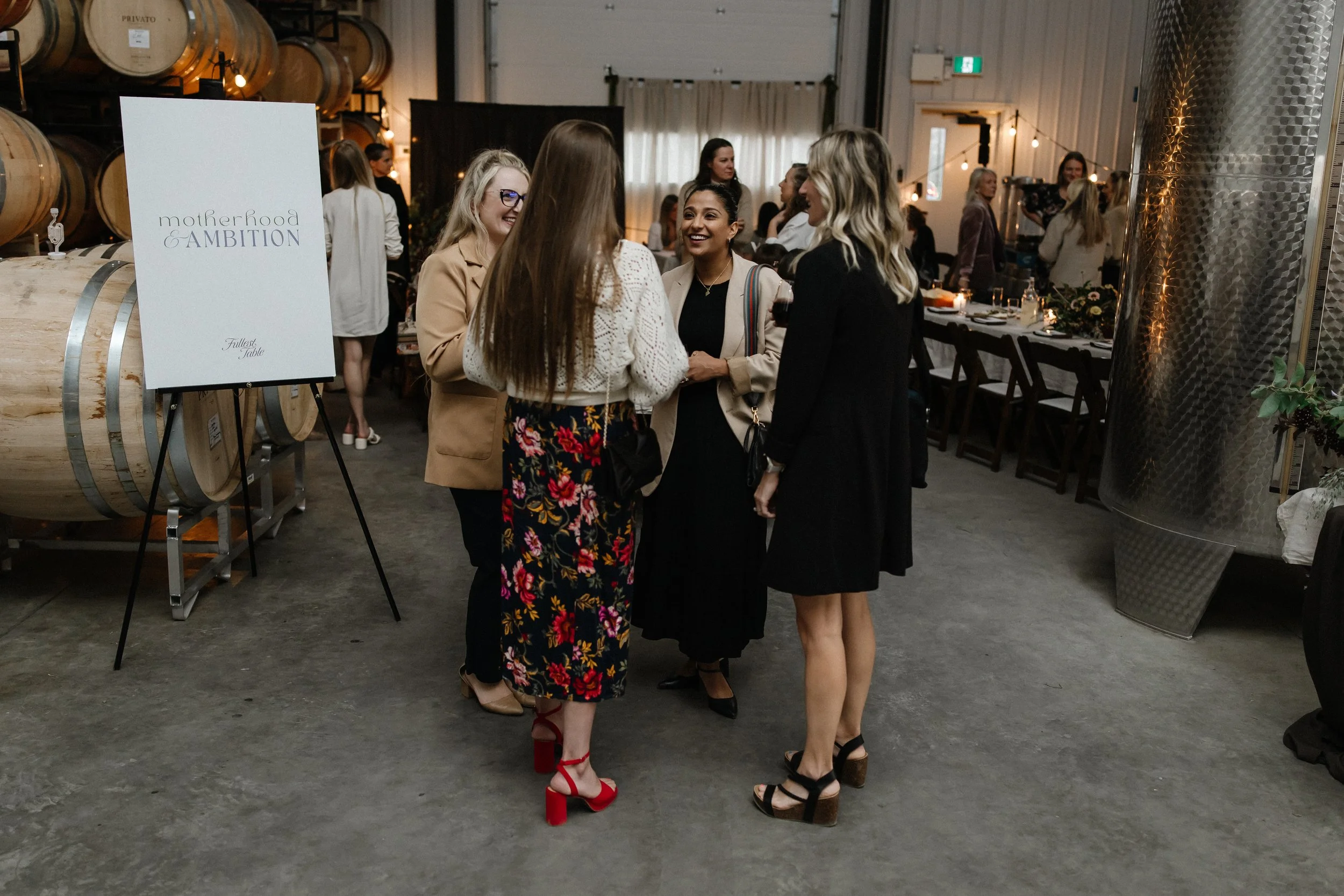 Group of women conversing at a social event inside a winery or brewery with barrels and a sign that reads 'motherhood & AMBITION' visible.