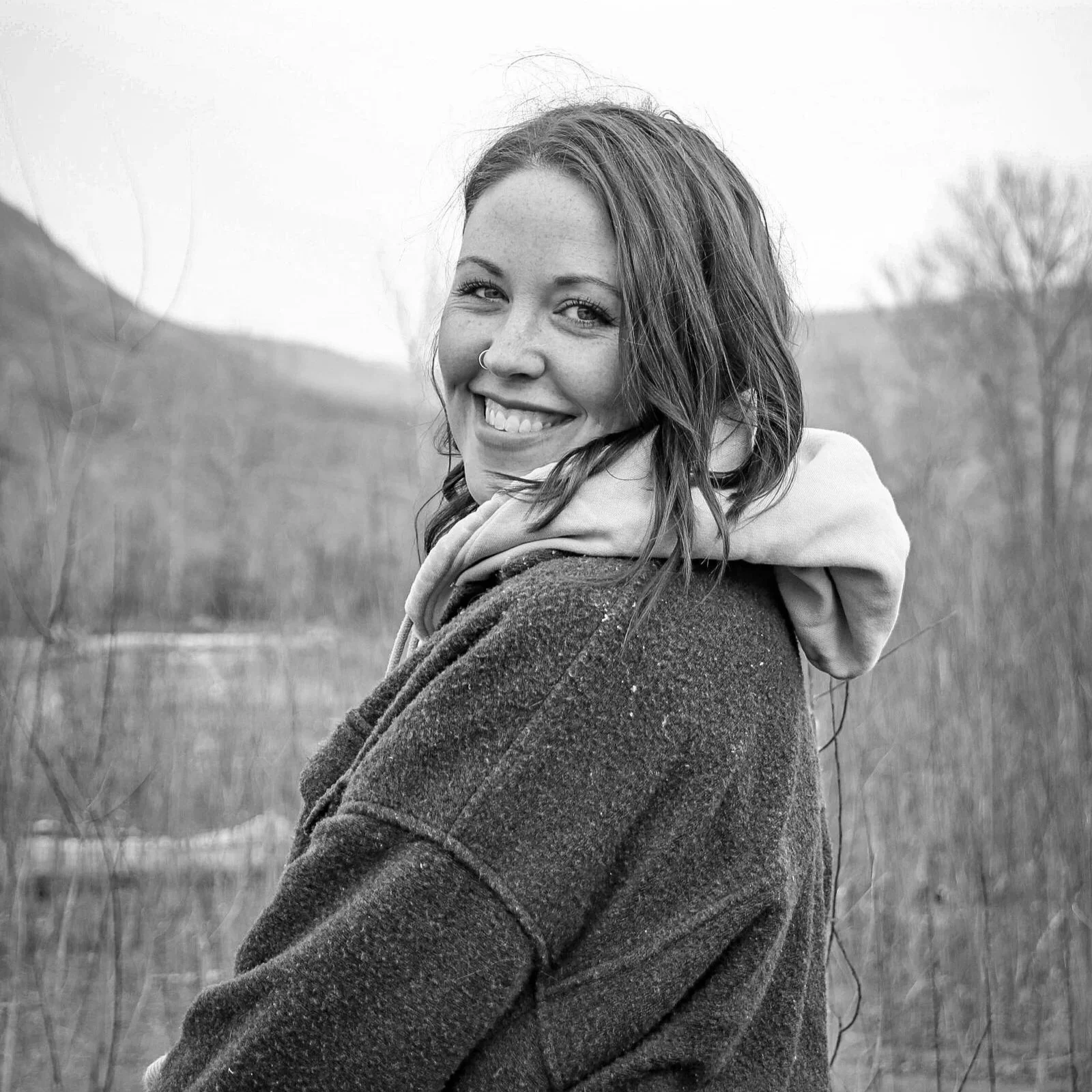 A smiling woman with wavy hair and a nose ring, wearing a hoodie under a jacket, standing outdoors in a natural setting with trees and hills in the background.