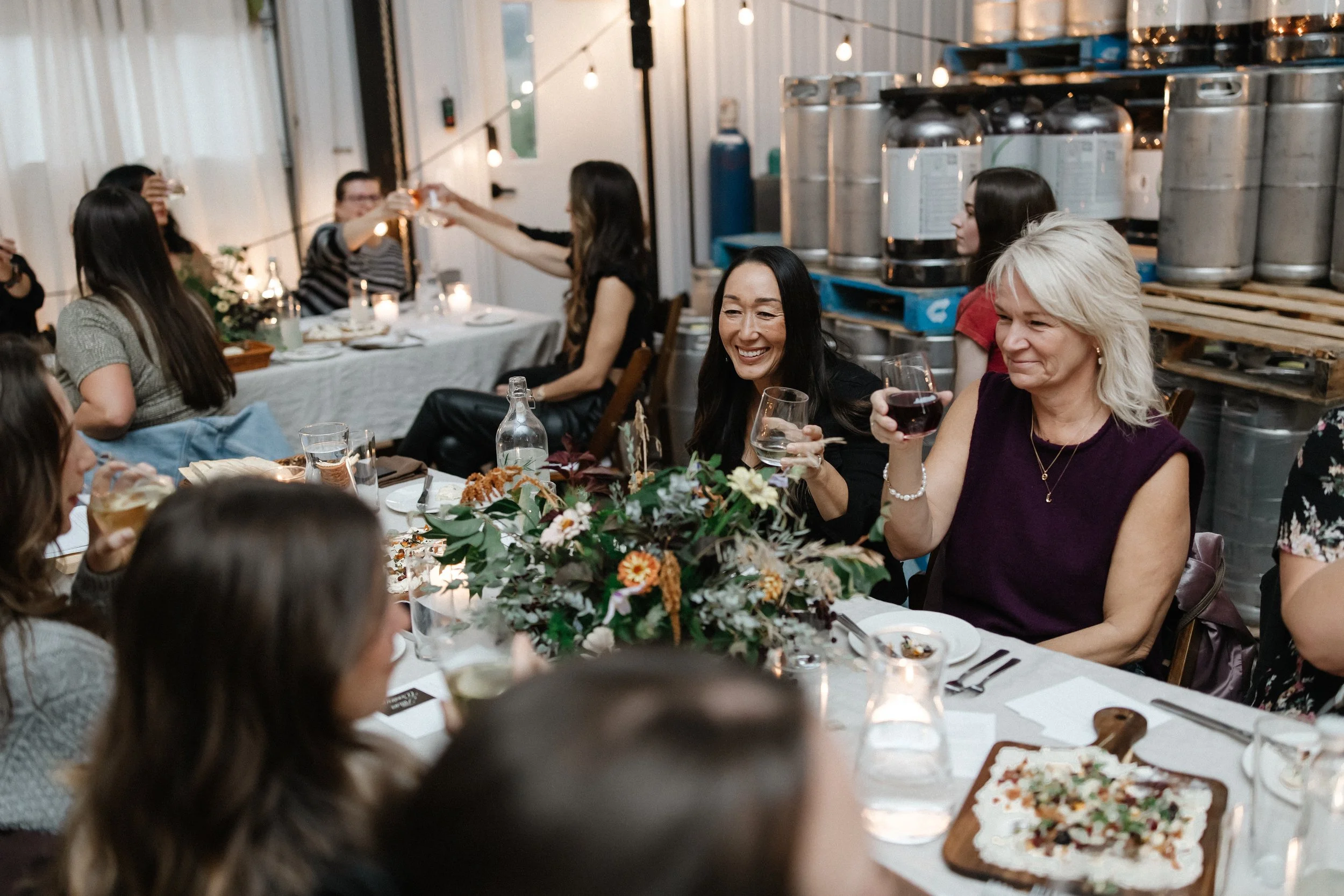 People gathered at a celebration dinner, raising glasses in a toast, with a floral centerpiece and food on the table, in a cozy, decorated setting.