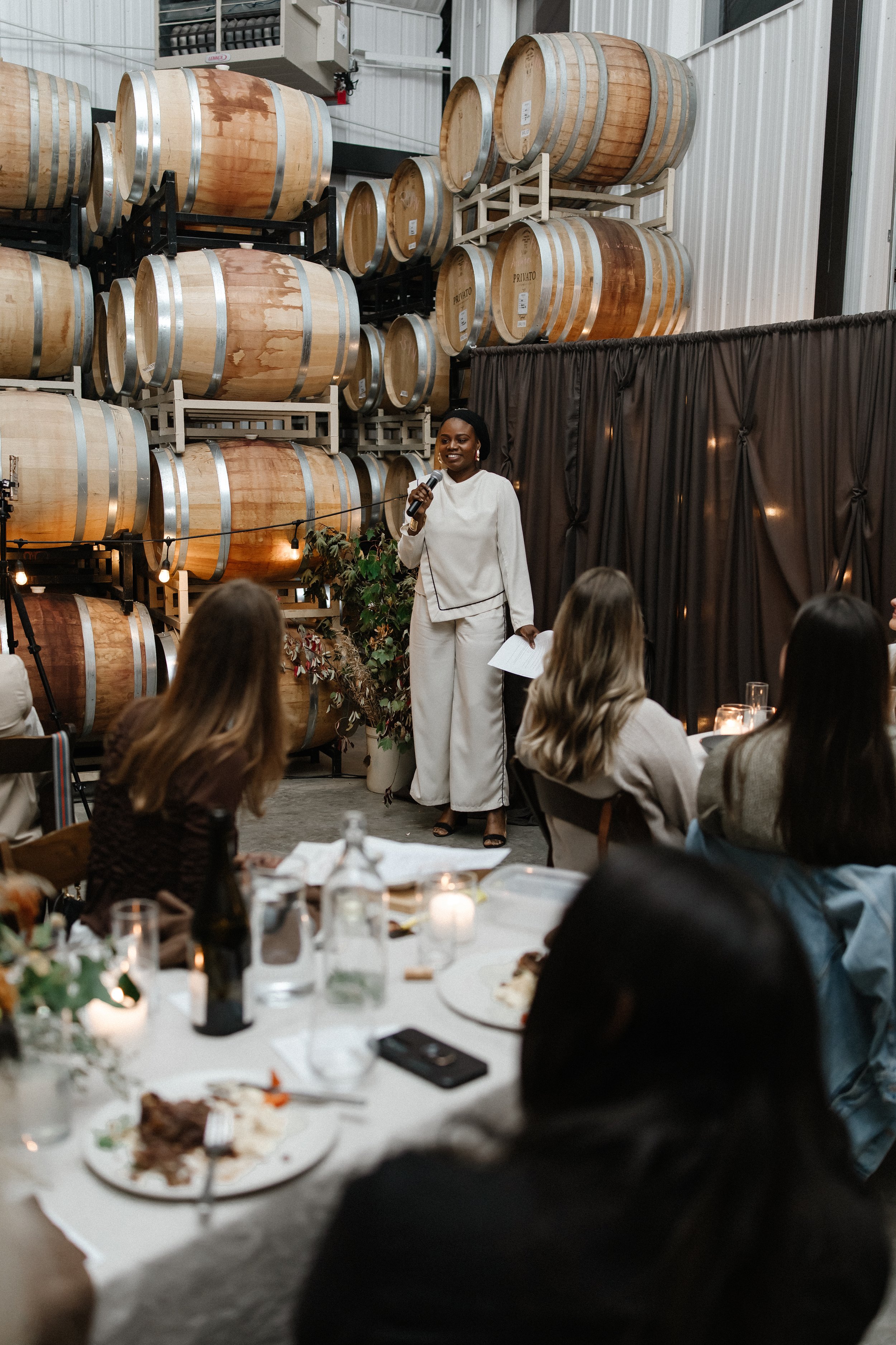 A woman in white standing on stage with a microphone, speaking to an audience in a winery or cellar with wine barrels stacked in the background.