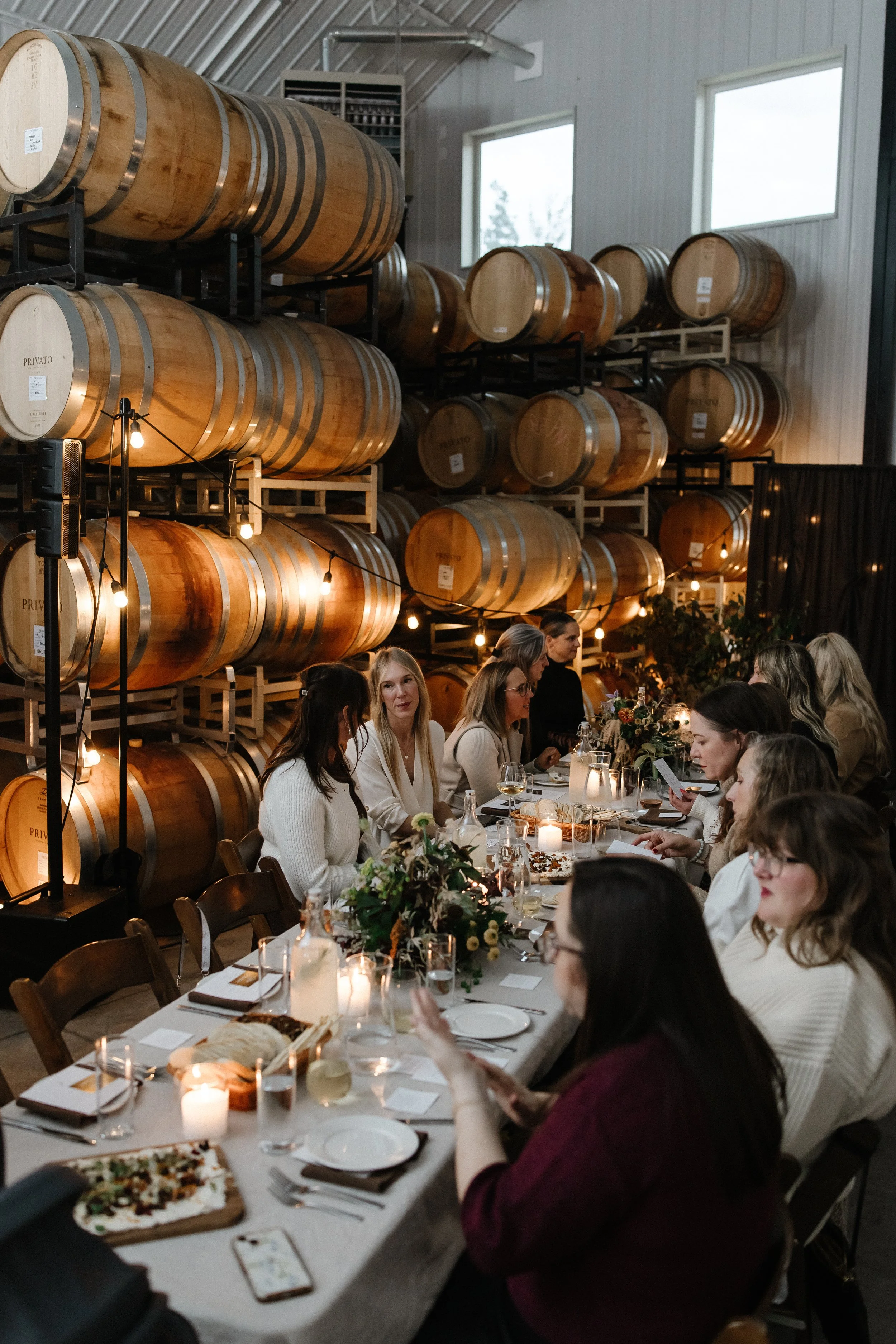 Group of women sitting at a long table in a winery tasting room with wine barrels in the background, decorated with candles and floral centerpieces.