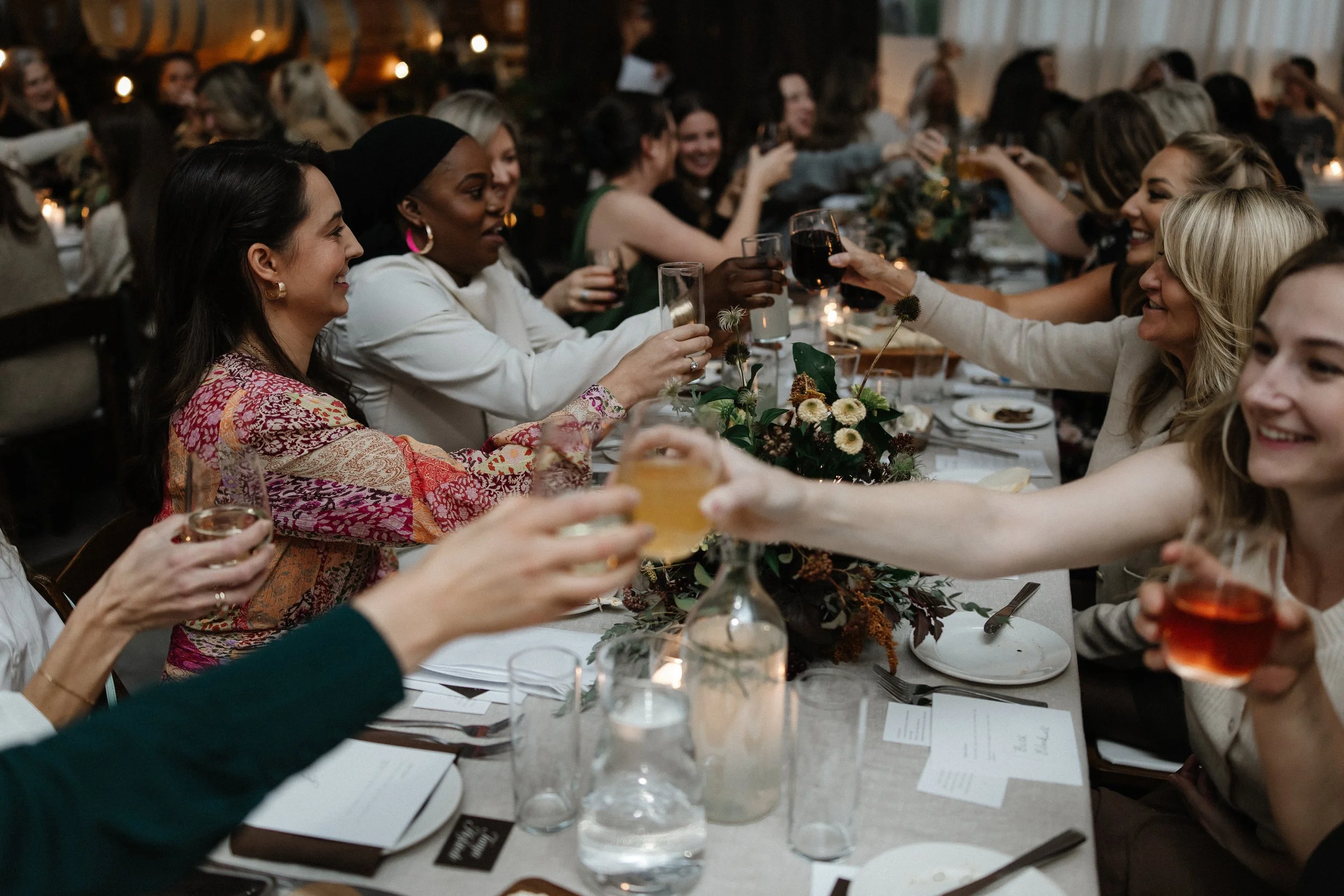 People sitting at a long dining table, raising glasses for a toast during a celebration or dinner event.