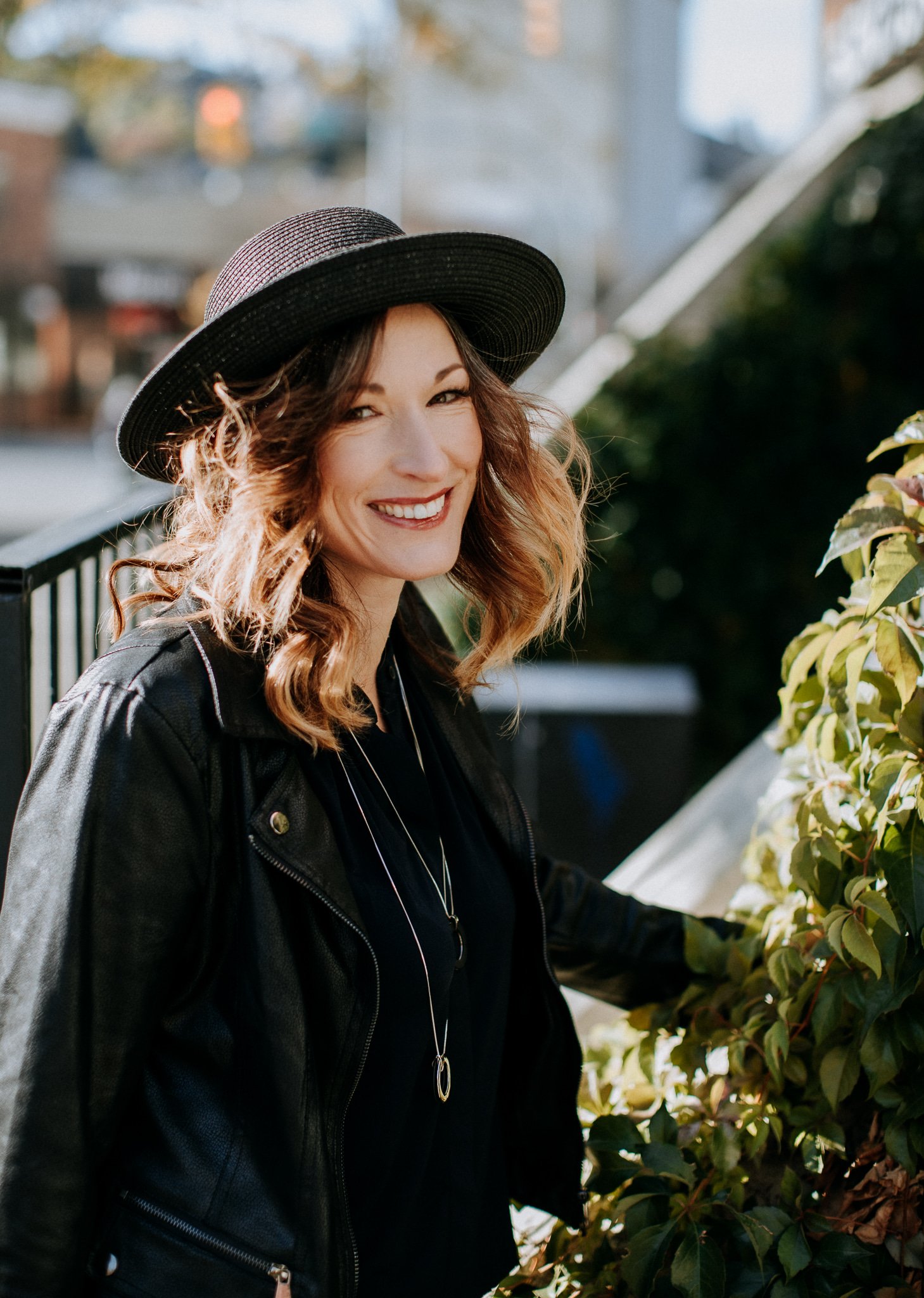 A woman with curly hair wearing a black hat and leather jacket outdoors, smiling at the camera, standing near green bushes.