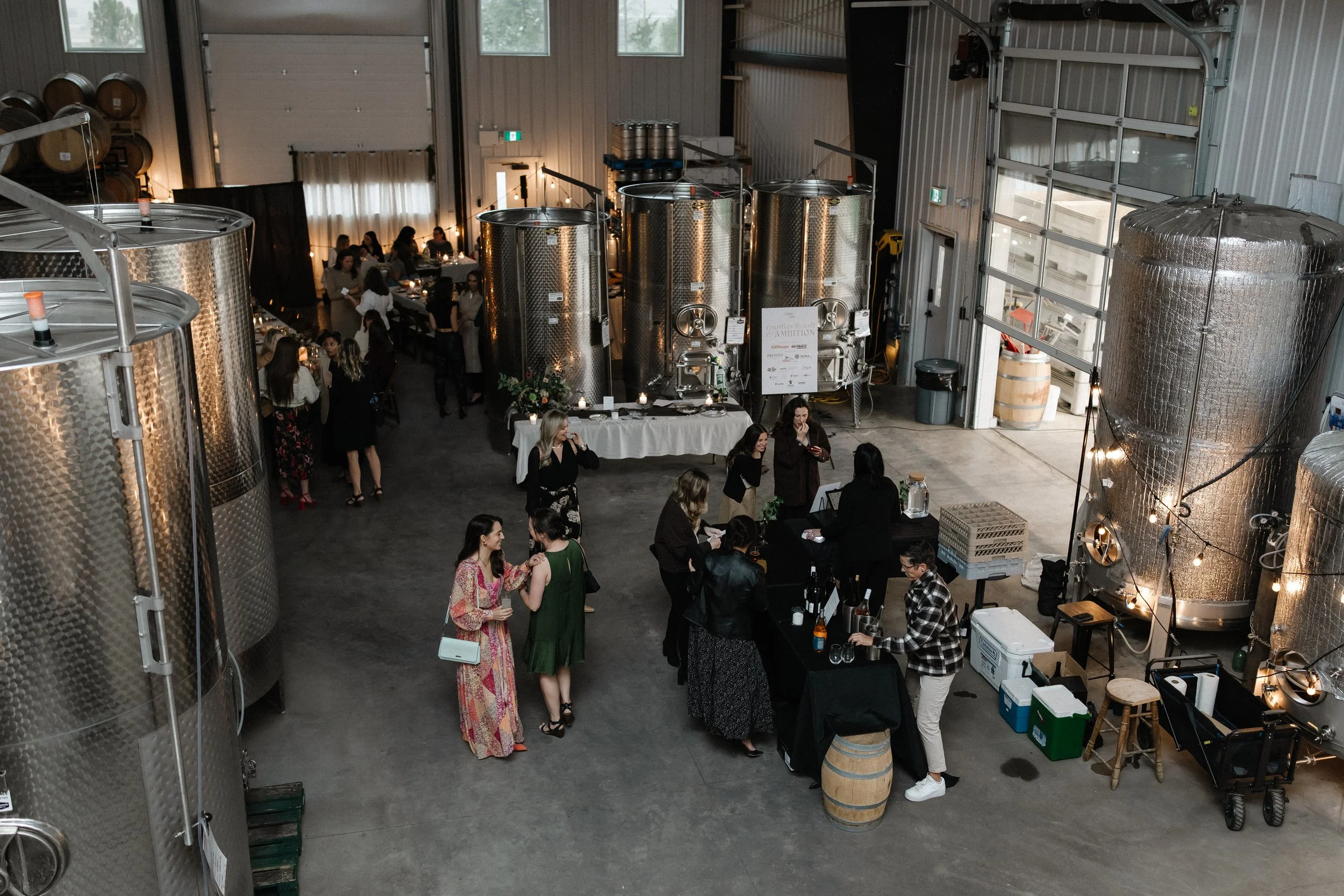 People socializing in a brewery with large fermentation tanks in the background and a bar area in the foreground.