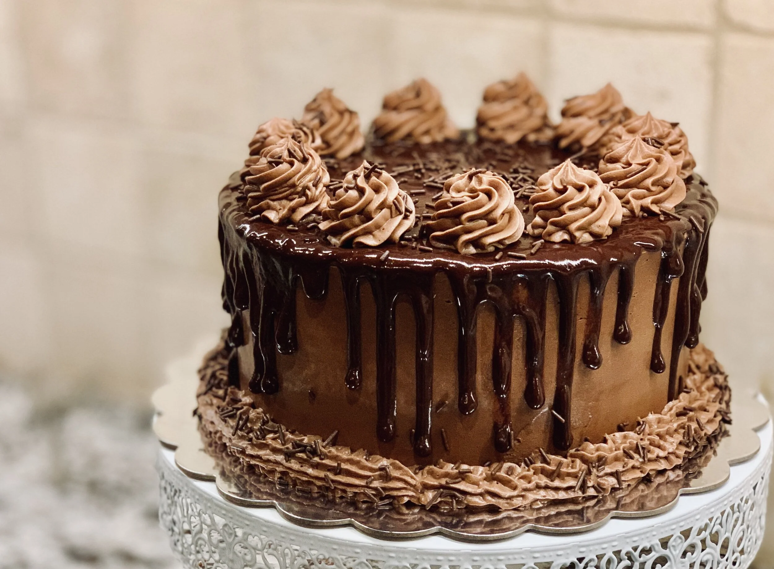 A chocolate cake with chocolate frosting, topped with piped chocolate whipped cream and chocolate sprinkles, with a glossy chocolate ganache drip on the sides, on a white decorative cake stand.