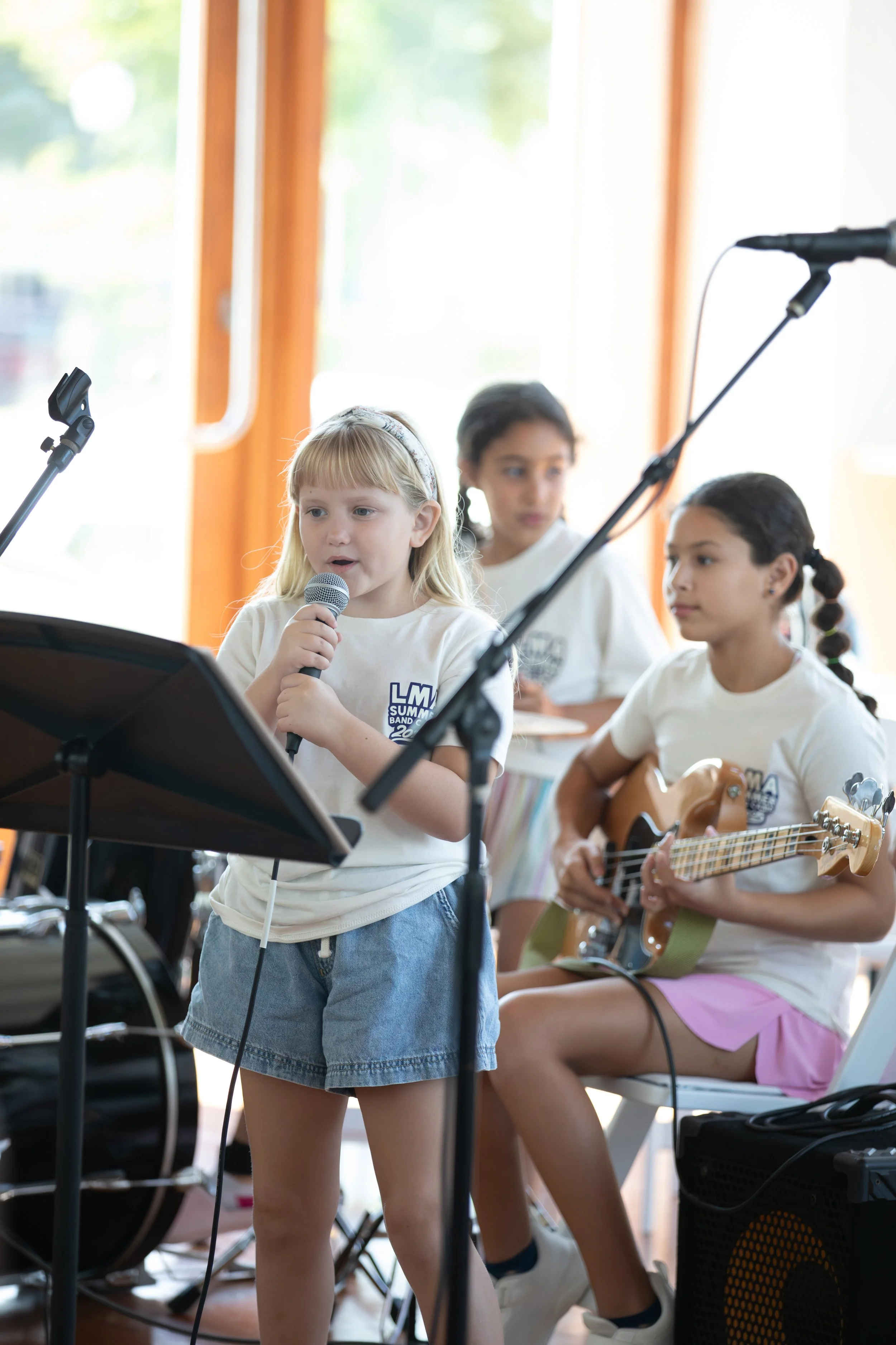 Young girls performing music together, one speaking into a microphone, another playing a guitar, in a bright indoor space at Summer Camp in Coppell Texas.