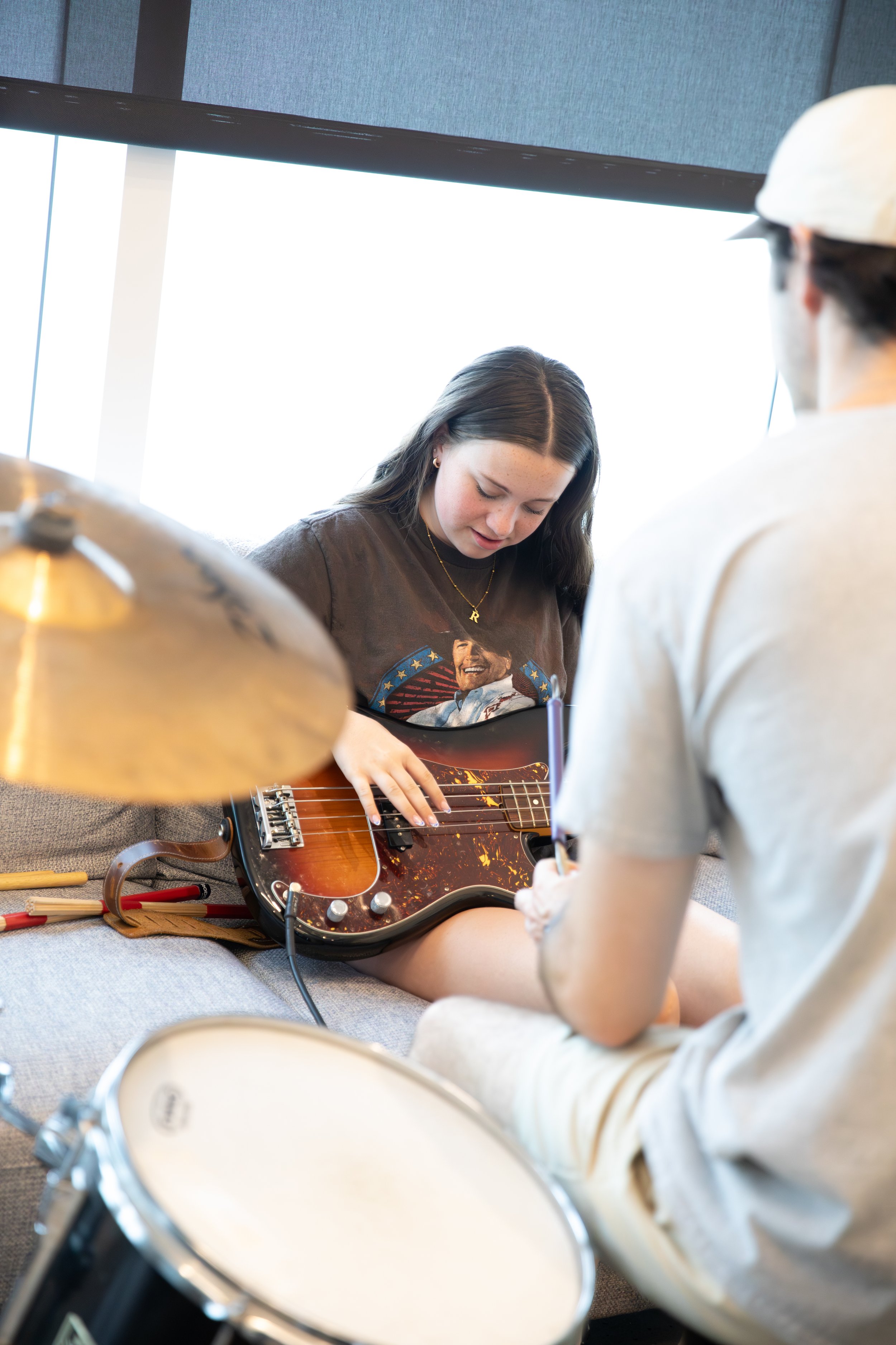 A young woman is sitting on a couch, playing an electric guitar while a person with a drumstick appears to be taking notes or writing, suggesting a music practice or lesson in a room with large windows at Summer Camp in Coppell Texas.