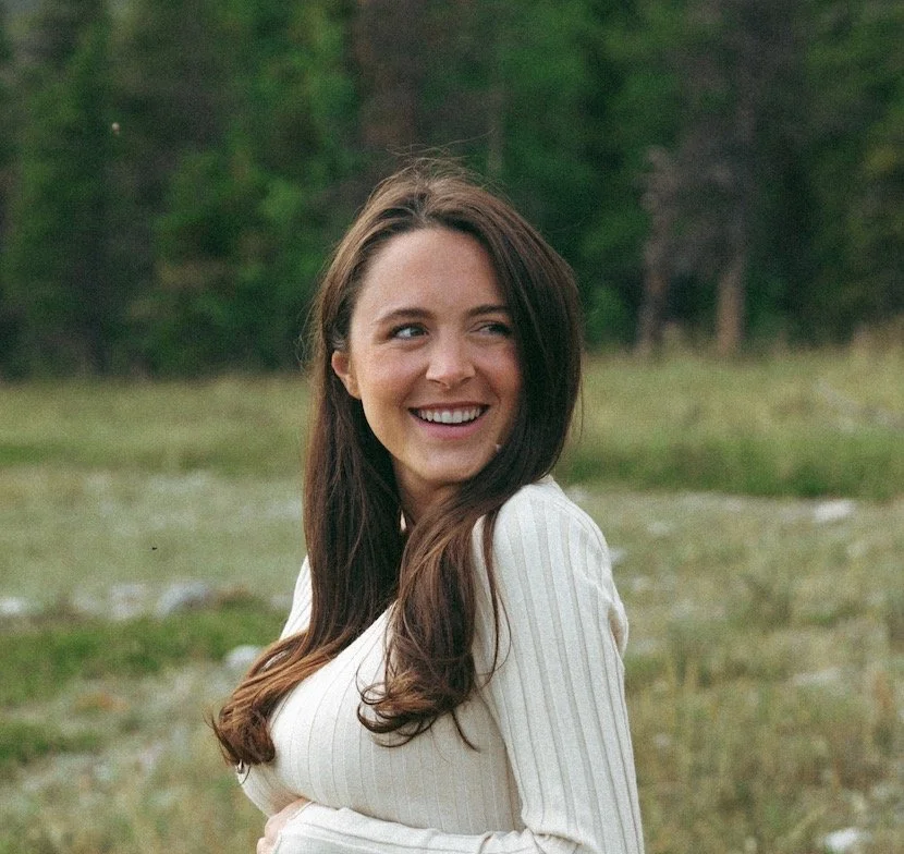 A woman with long brown hair, wearing a white ribbed sweater, smiling outdoors in a grassy, wooded area.