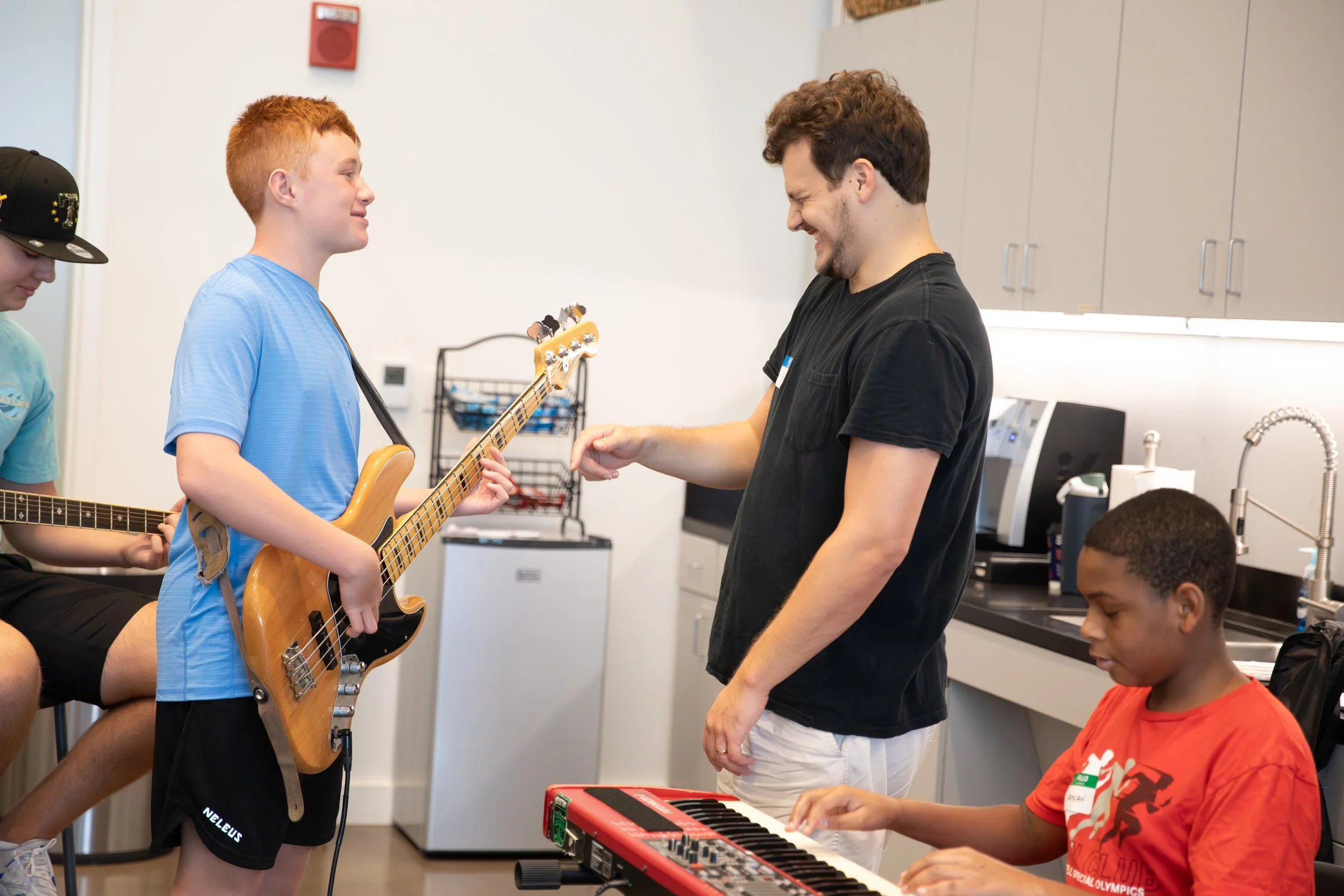 A group of kids with a man in a room, some playing musical instruments and the man smiling. Kid in blue shirt playing bass guitar, boy in red shirt playing keyboard, and another kid partially visible with guitar at Summer Camp in Coppell Texas.