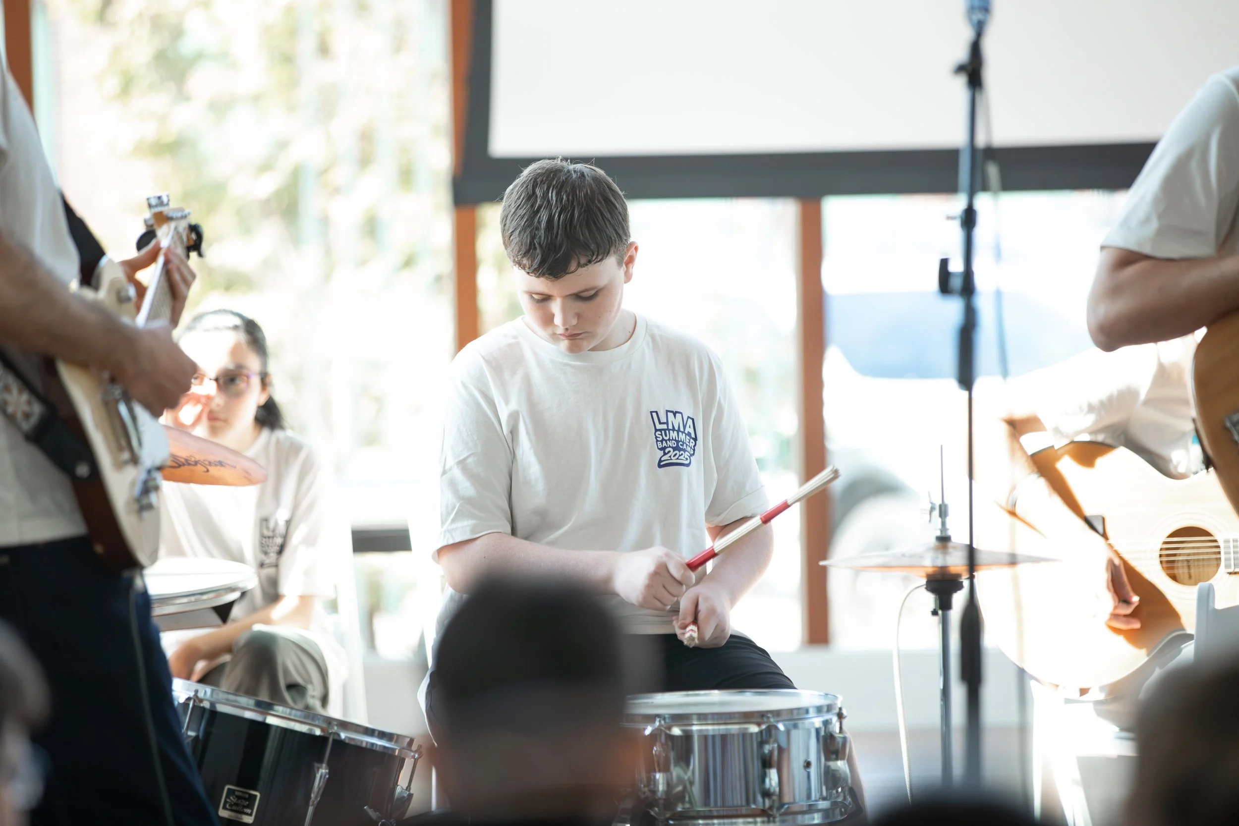 Young boy playing drums during band practice with other musicians in the background, including a girl with glasses and a person playing guitar at Summer Camp in Coppell Texas.