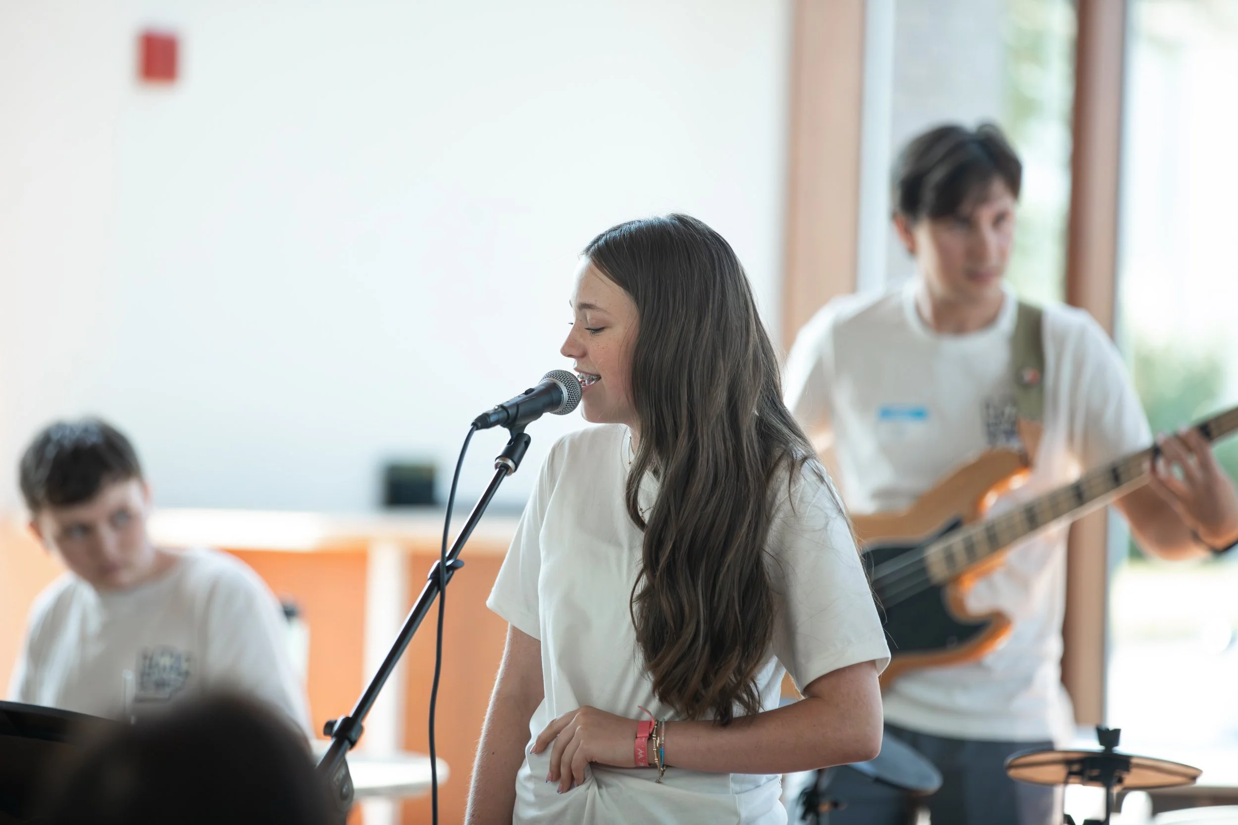 Girl singing into a microphone while two boys play instruments in the background during a musical performance at Summer Camp in Coppell Texas.