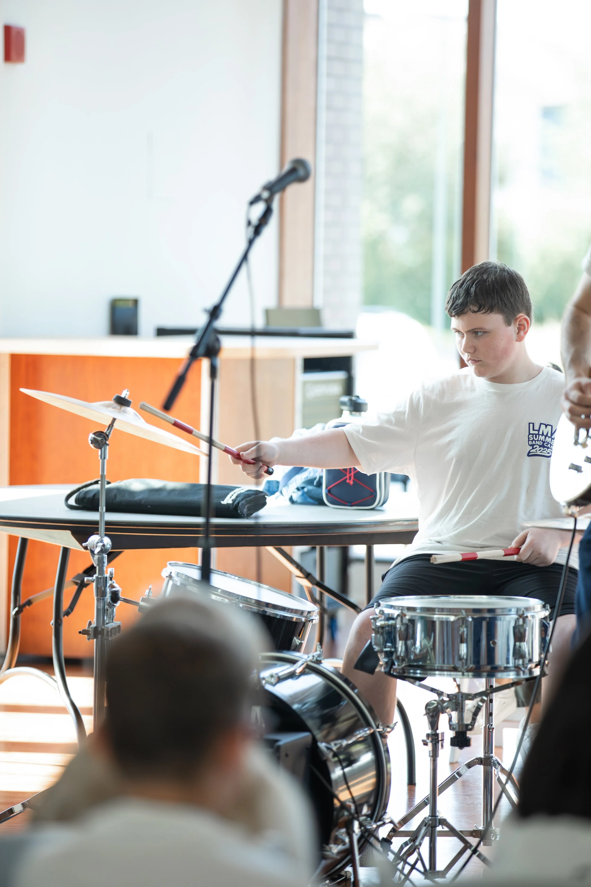 Young boy playing drums during a band practice session in a bright room with large windows at Summer Camp in Coppell Texas.