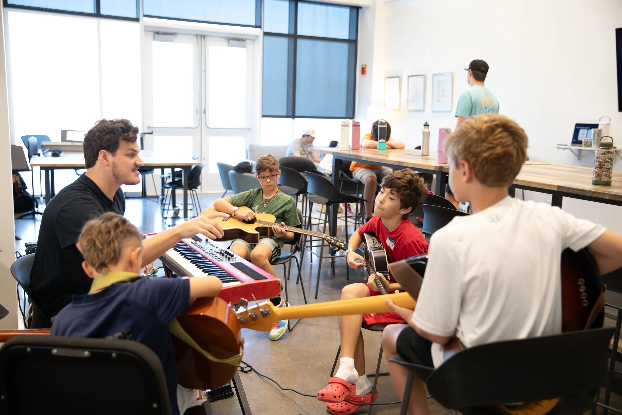 A group of children and a young adult in a music classroom, playing guitars and keyboard with a teacher. Other children are sitting at a table in the background at Summer Camp in Coppell Texas.