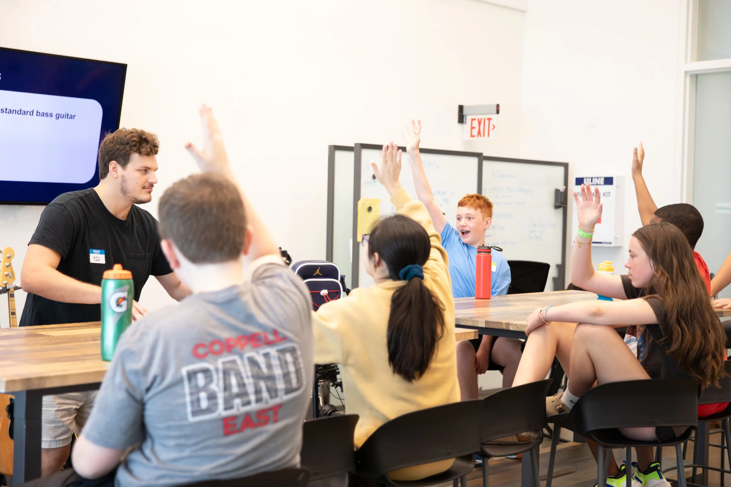 A classroom setting with a teacher and students, some students raising their hands, in a room with white walls, a wall-mounted monitor, and a whiteboard at Summer Camp in Coppell Texas.