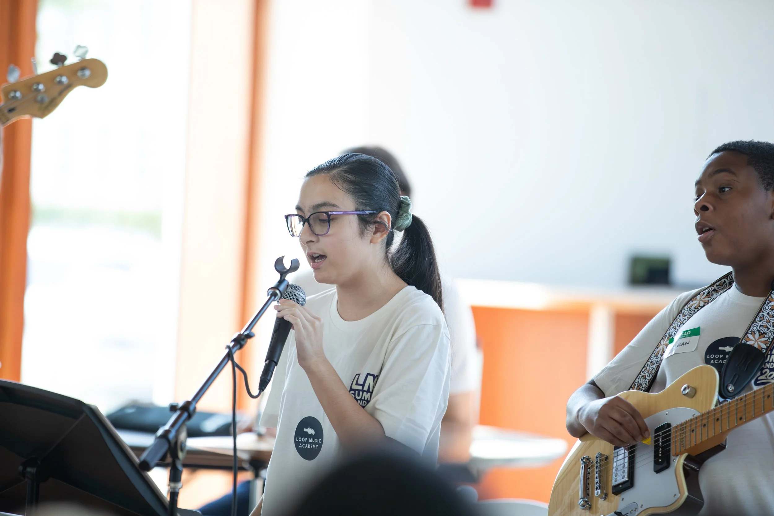 Young girl singing into a microphone while playing an electric guitar, with a young boy beside her also playing an electric guitar, in a music practice or performance setting at Summer Camp in Coppell Texas.