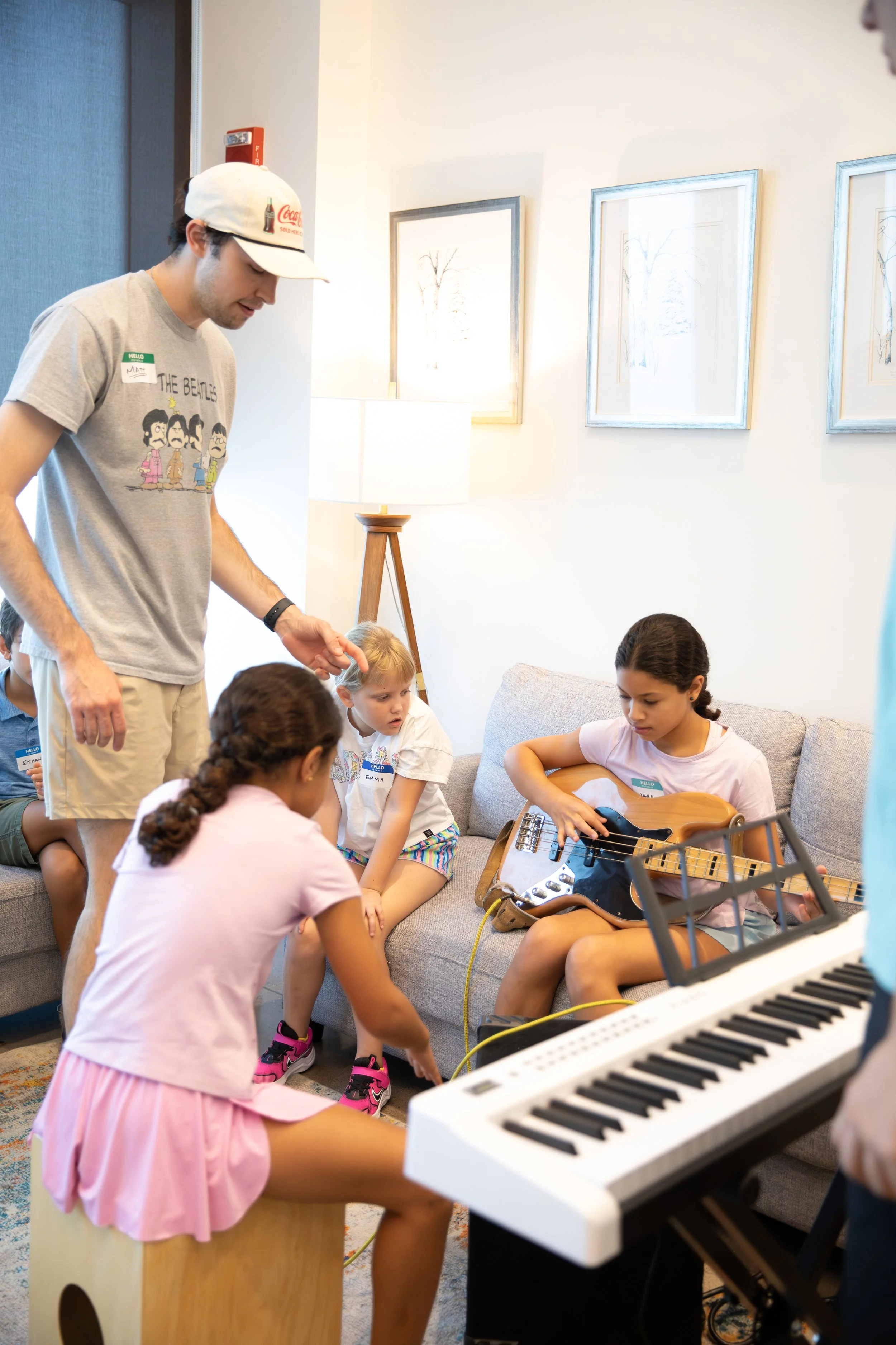 Children sitting on a couch, one playing guitar, another sitting on a stool, with a keyboard in the foreground, adults observing in a cozy room with framed art on the white wall at Summer Camp in Coppell Texas.