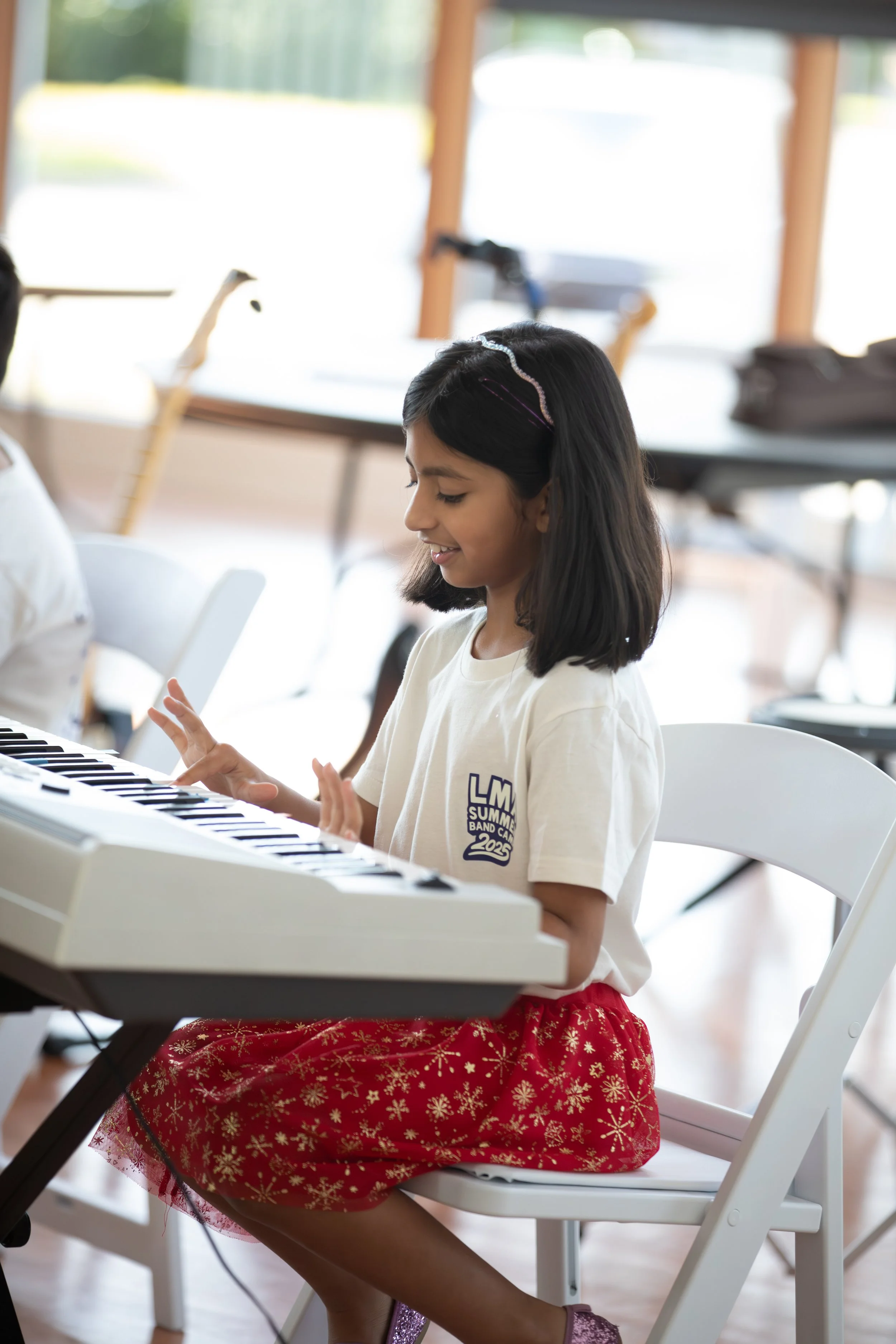 Young girl playing a keyboard, sitting on a white chair in a brightly lit room, wearing a white t-shirt with a logo, a red skirt with gold snowflake patterns, and sparkly shoes at Summer Camp in Coppell Texas.