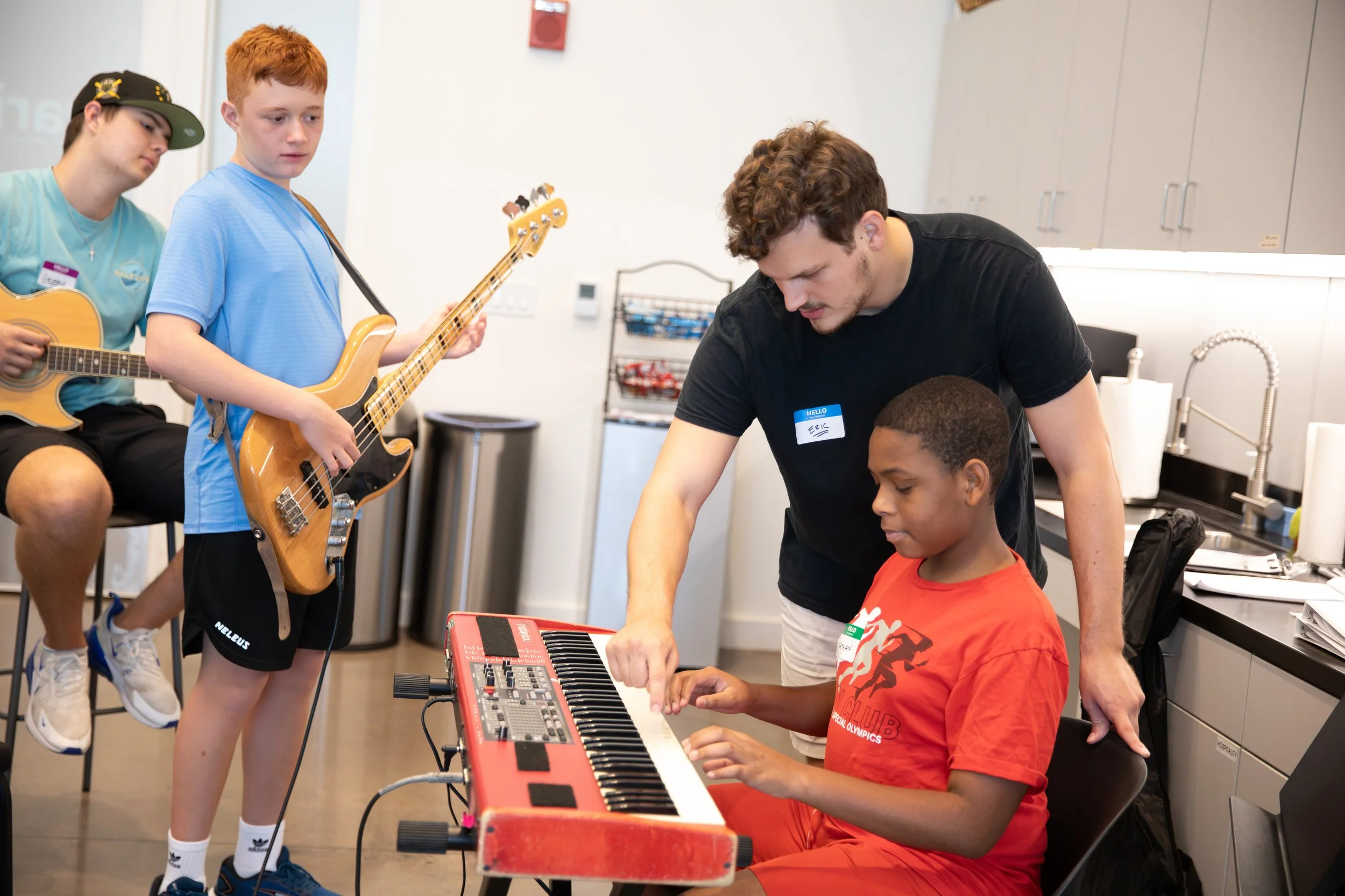 A music class with two kids playing guitars and one boy playing a keyboard, assisted by an instructor in a classroom or practice room at Summer Camp in Coppell Texas.
