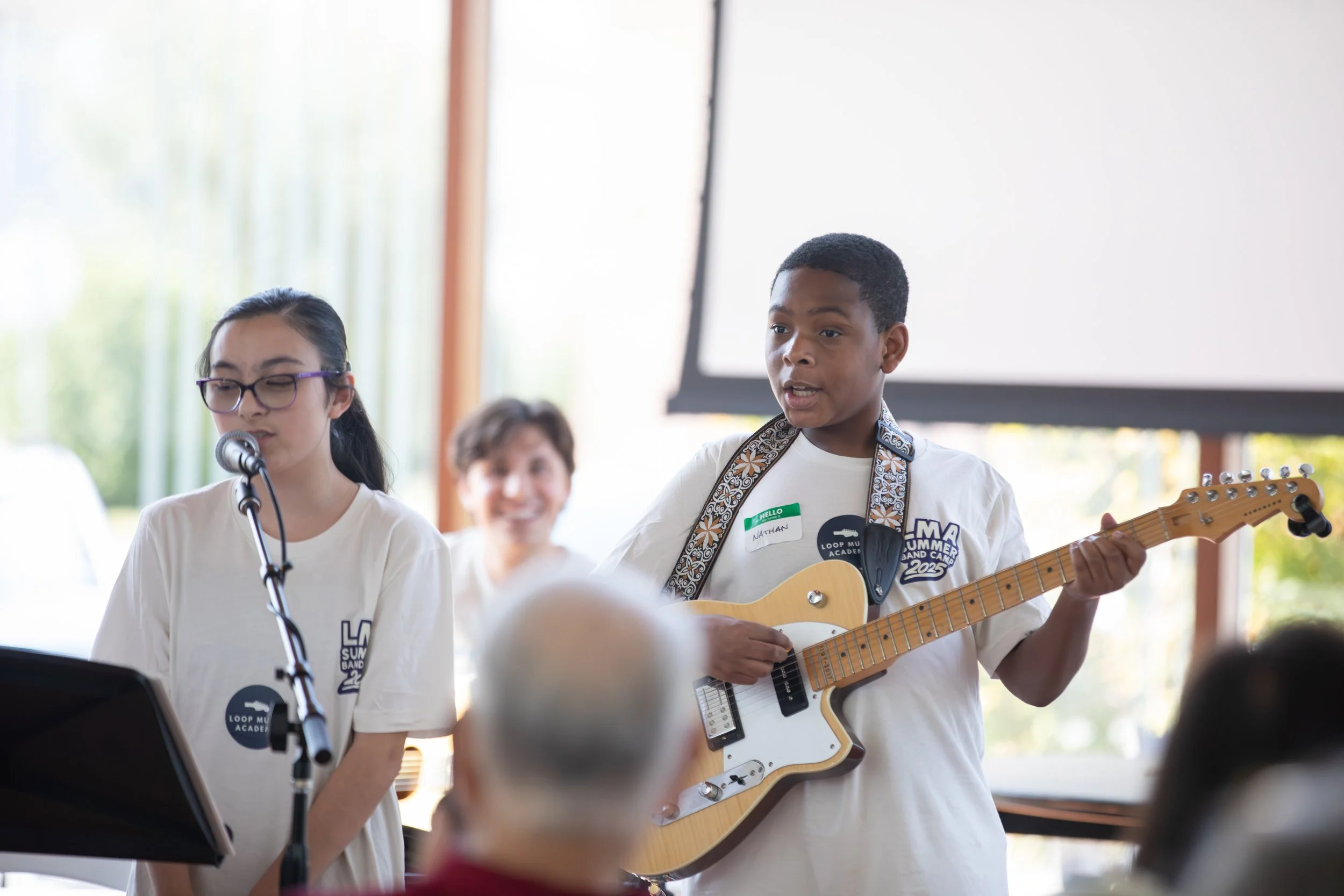 Young boy playing an electric guitar and singing into a microphone at a music camp event, with a young girl and an adult woman in the background, all in a bright room with large windows at Summer Camp in Coppell Texas.