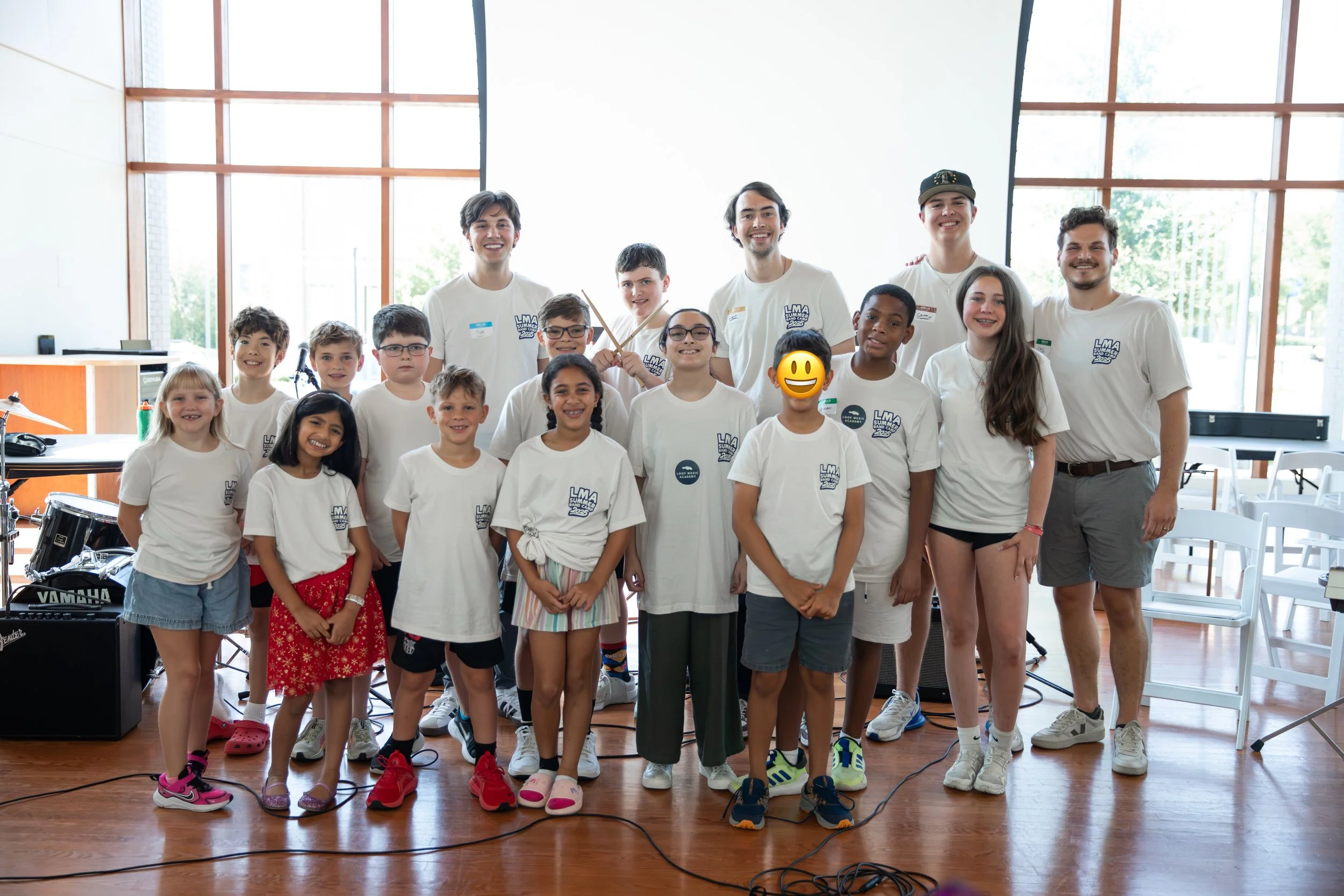 Group of children and young adults in a brightly lit room with large windows, smiling and posing for a photo, some wearing white t-shirts with a logo, musical instruments and equipment in the background.