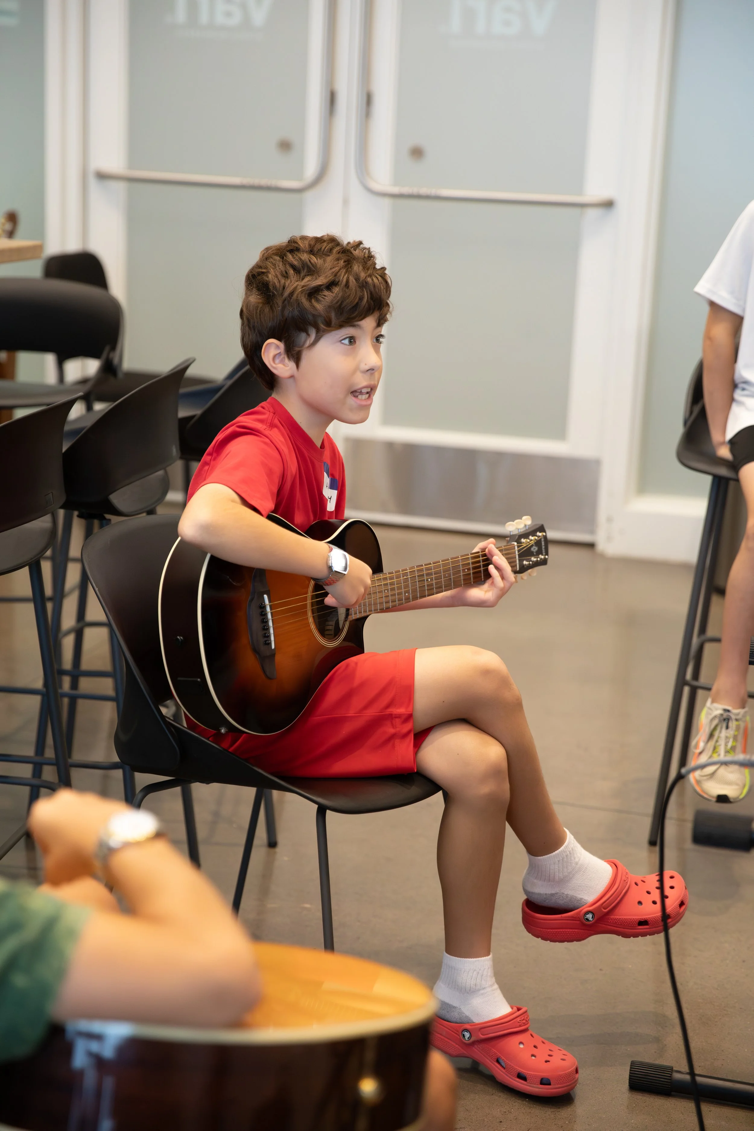 A young boy with curly brown hair sitting on a black chair, playing an acoustic guitar in a room with other chairs and a closed door in the background at Summer Camp in Coppell Texas.