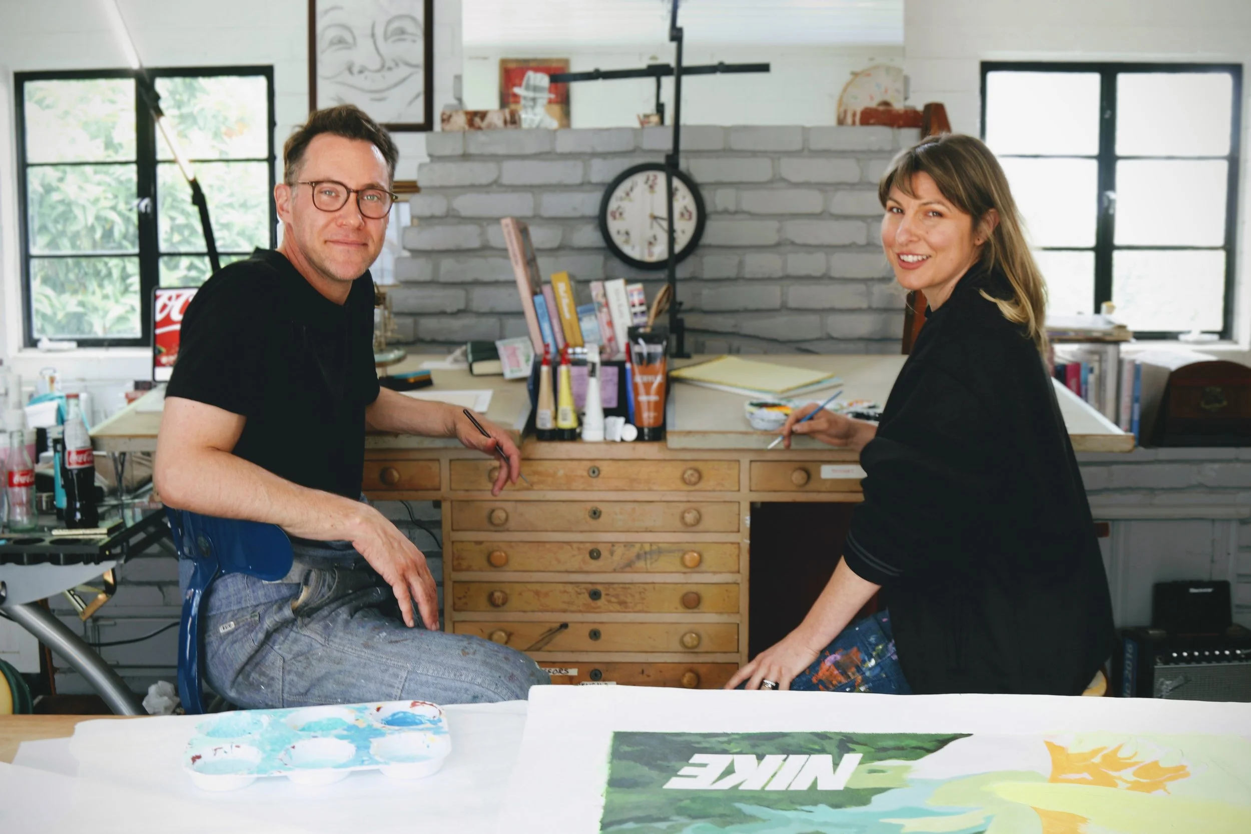 Two people, a man and a woman, are seated at a work table in an art studio, smiling and painting. The studio has large windows, a gray brick wall, and art supplies in the background.