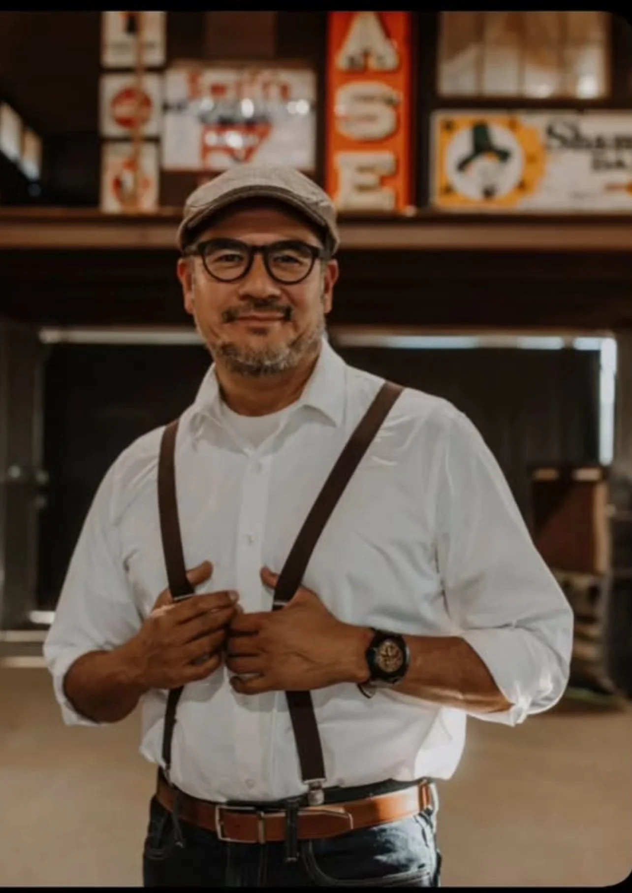 A smiling man wearing glasses, a flat cap, a white shirt, suspenders, and a wristwatch, standing indoors with a blurred background that includes signs and decorations.