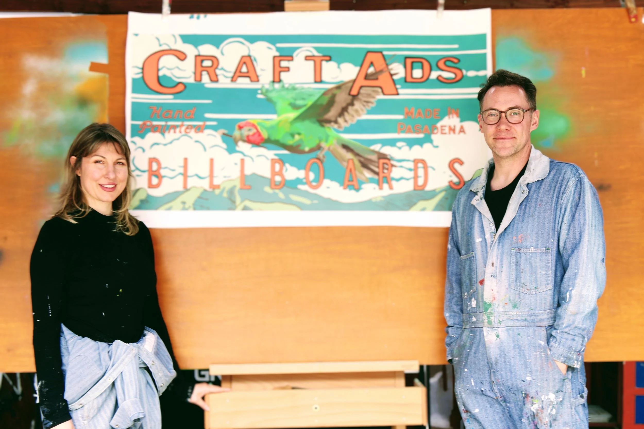 A woman and a man standing in front of a colorful sign that reads 'Craft Ads Hand Painted Made in Pasadena Billboards'. The woman wears a black top and has a gray jacket tied around her waist. The man wears glasses and is dressed in paint-splattered overalls. They are smiling.