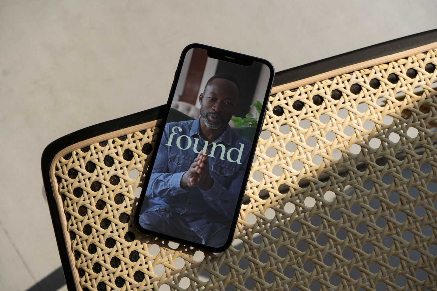 Smartphone with an image of a man in a denim shirt and the word 'found' on the screen, resting on a woven rattan surface.