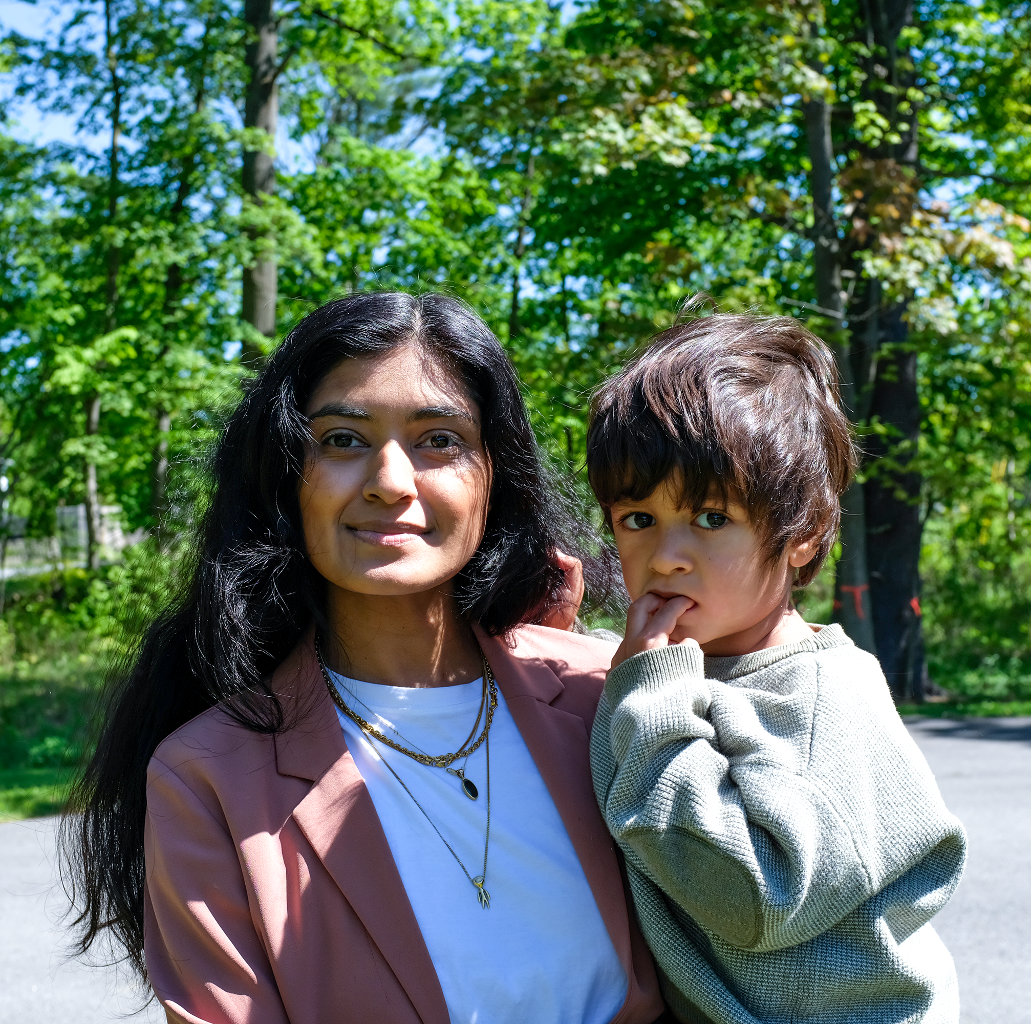 A woman holding a young boy outdoors on a sunny day with green trees in the background.