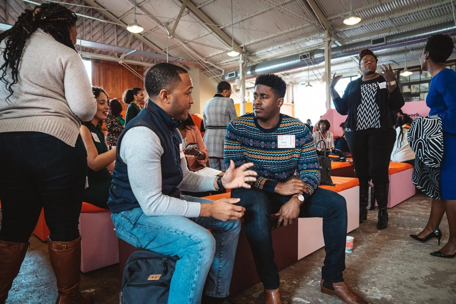 A group of diverse people are sitting and standing in a conversation at a networking event or conference in an indoor space with exposed ceiling pipes and industrial lighting.