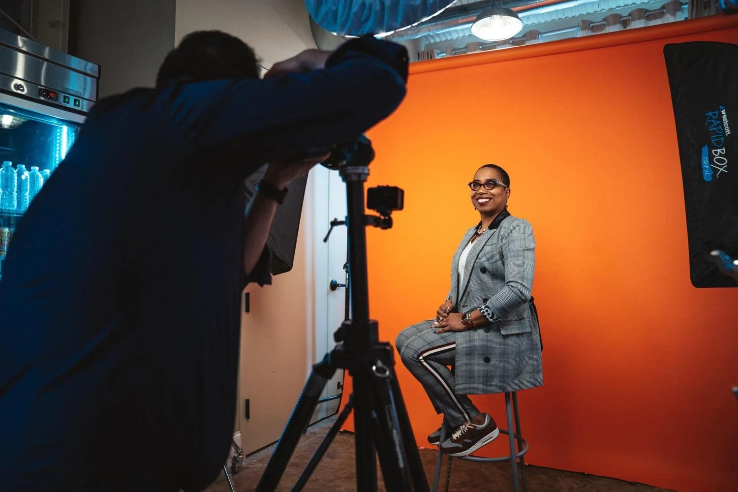 Woman in a plaid blazer sitting on a stool and smiling in front of an orange backdrop during a photo shoot.