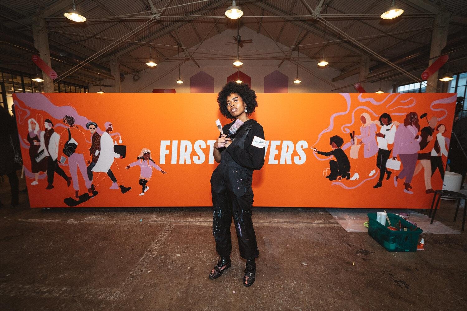 A woman standing in front of a large orange mural with text that reads 'FIRST' and illustrations of diverse people walking and taking photos.
