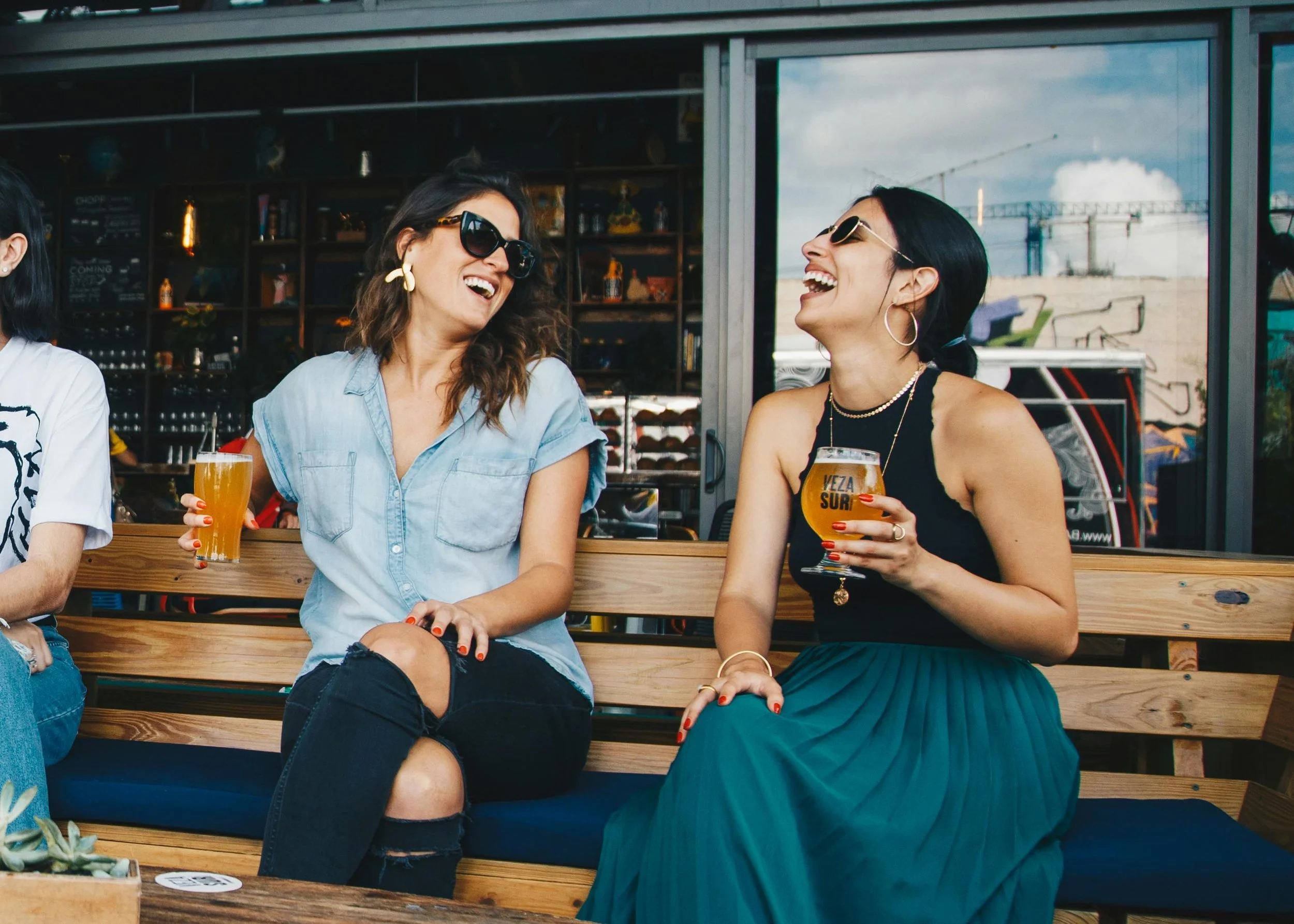 Two women sitting on a wooden bench, laughing and enjoying drinks outdoors, with a glass of beer and cocktail, sunglasses, and casual attire.