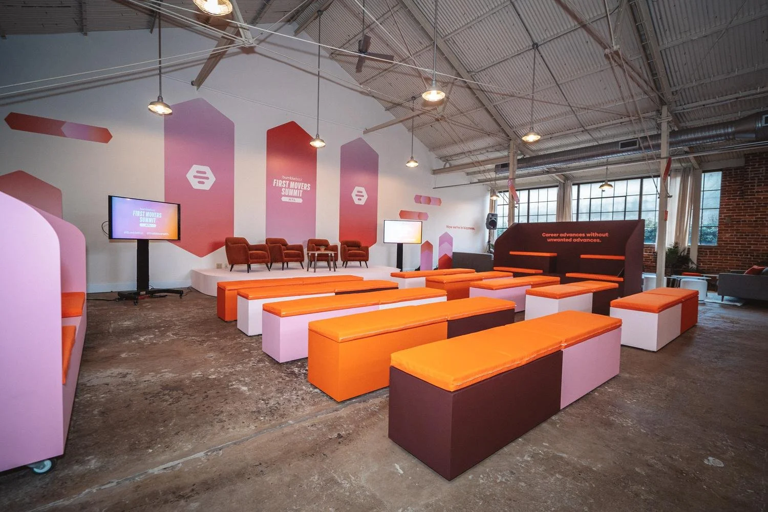 Conference room with orange and pink seating, stage with chairs, and large windows in an industrial-style building.