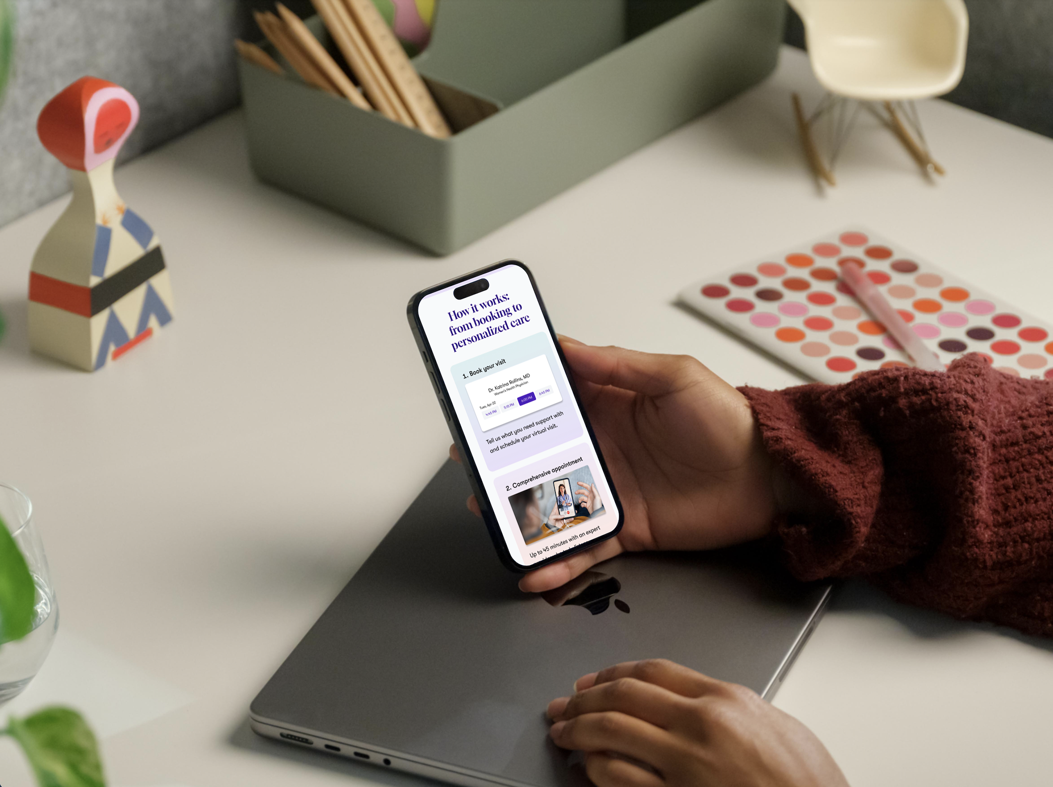 A person wearing a maroon sweater holding an iPhone displaying a booking website, with a closed laptop underneath, on a white desk alongside various decorative items and a pink eyeshadow palette.