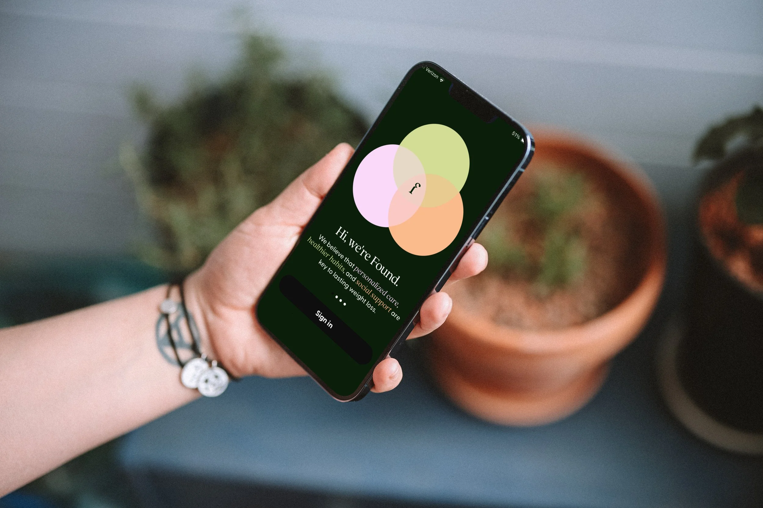 Person holding a smartphone with an app displayed on the screen, showing a colorful Venn diagram and welcoming message about health habits, with potted plants in the background.