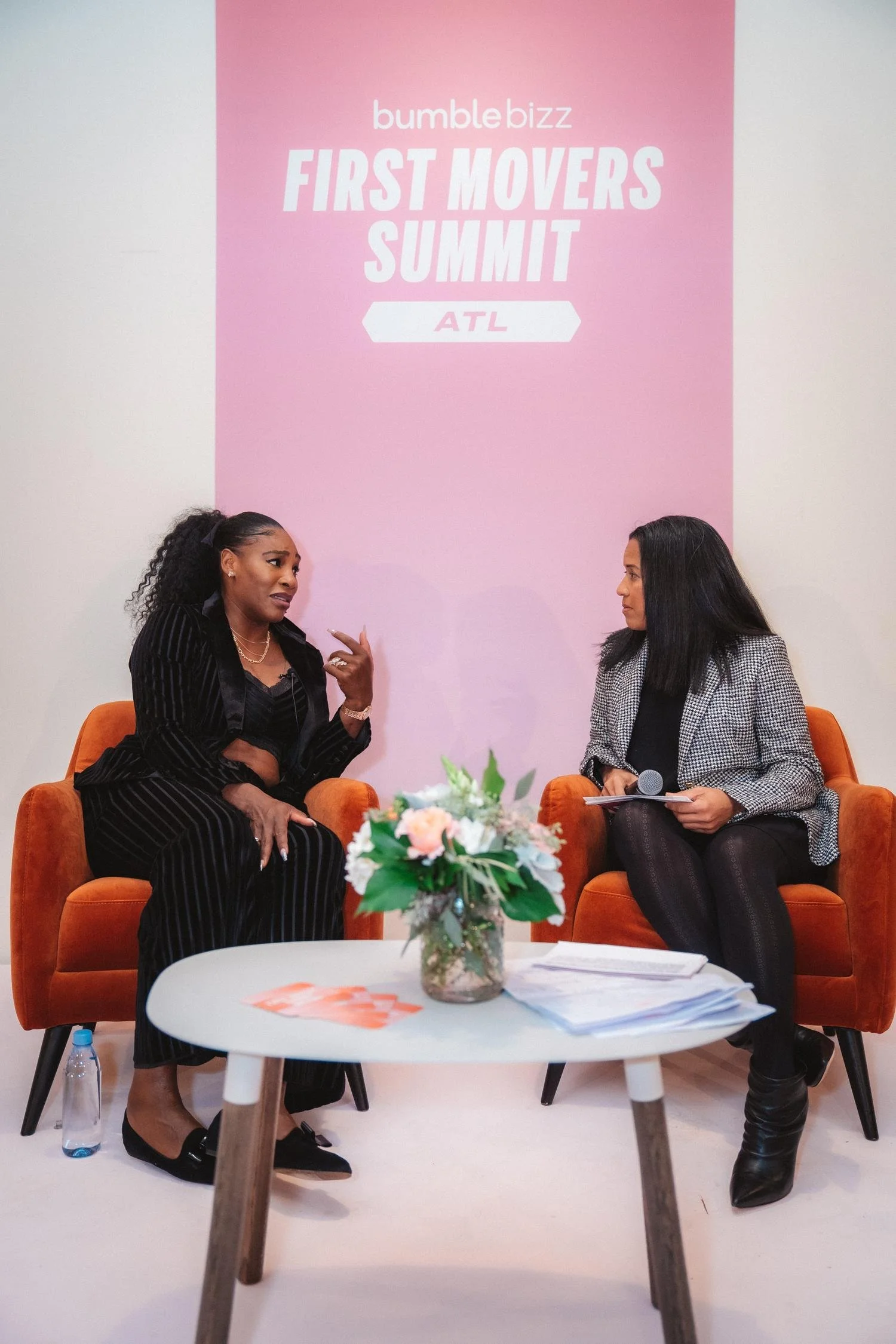 Two women sitting on orange armchairs having a conversation at the Bumble Bizz First Movers Summit in Atlanta, GA, with a pink backdrop and a flower arrangement on a white coffee table in front of them.