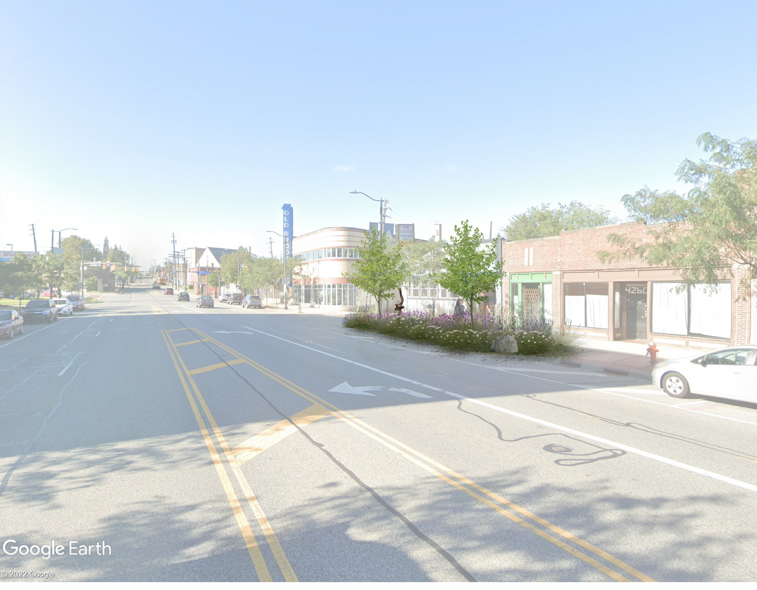 A small town street scene on a sunny day, with a wide road, some parked cars, trees, and view of buildings including a vintage-style theater with a blue sign, and storefronts along the sidewalk.