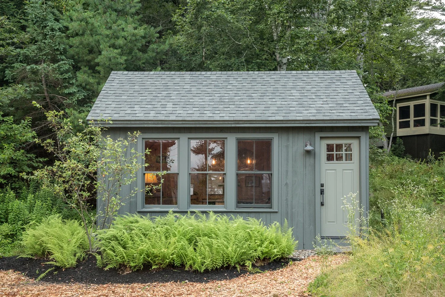 Small gray house with large windows and front door, surrounded by trees and green plants.