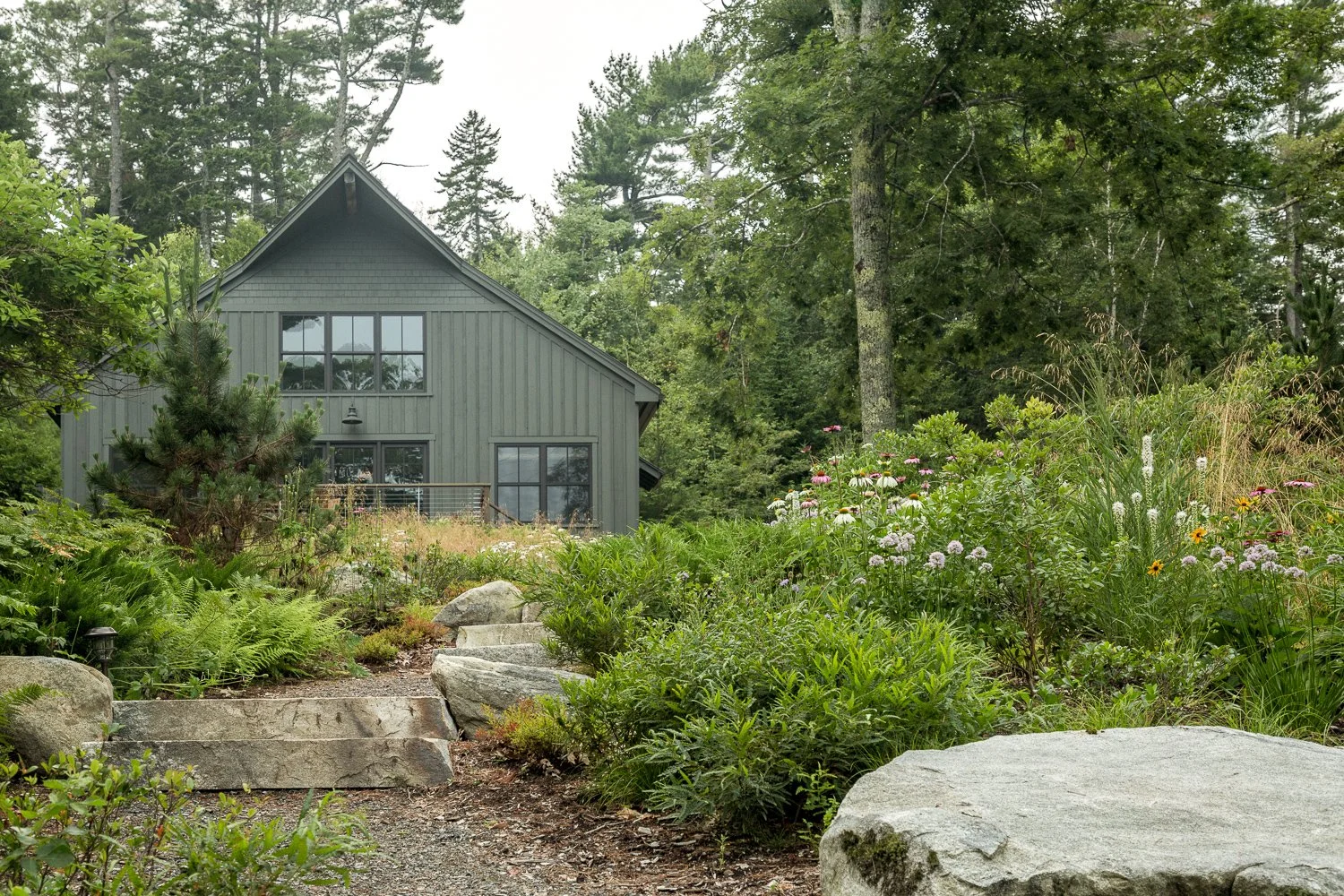 A gray house surrounded by lush greenery, trees, and wildflowers with stone steps leading up to the house.