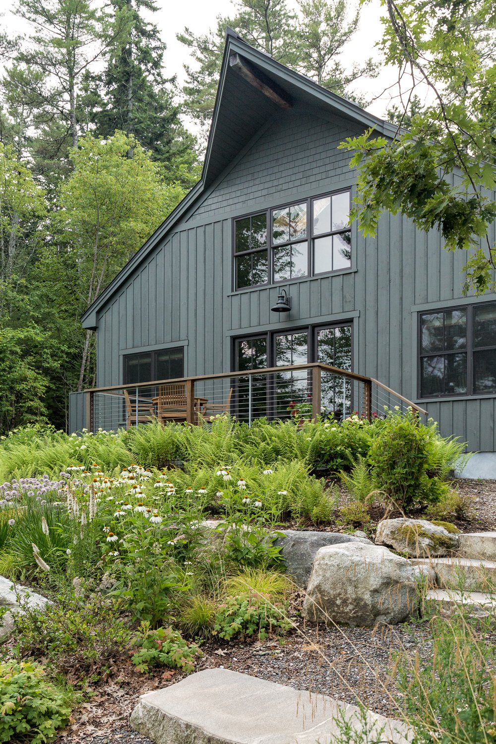 A modern house with blue-gray exterior siding, large windows, and a wooden deck with railing, surrounded by lush green plants and trees.