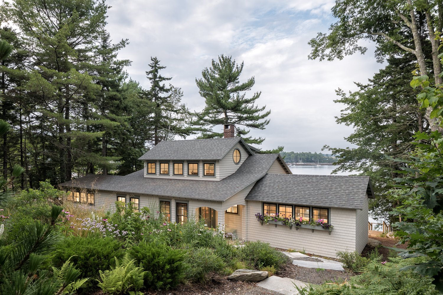 A modern house with multiple rooflines surrounded by lush greenery, overlooking a body of water in the background. The house has large windows with flower boxes.