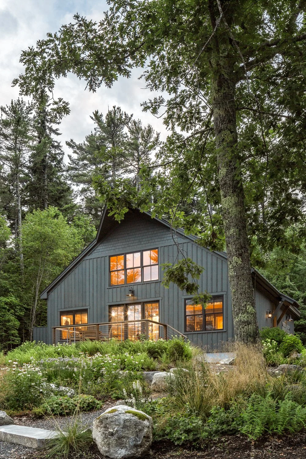 A modern dark blue house with large windows and a wooden balcony sitting in a lush green forest with trees and plants, sunset reflections visible in the windows.