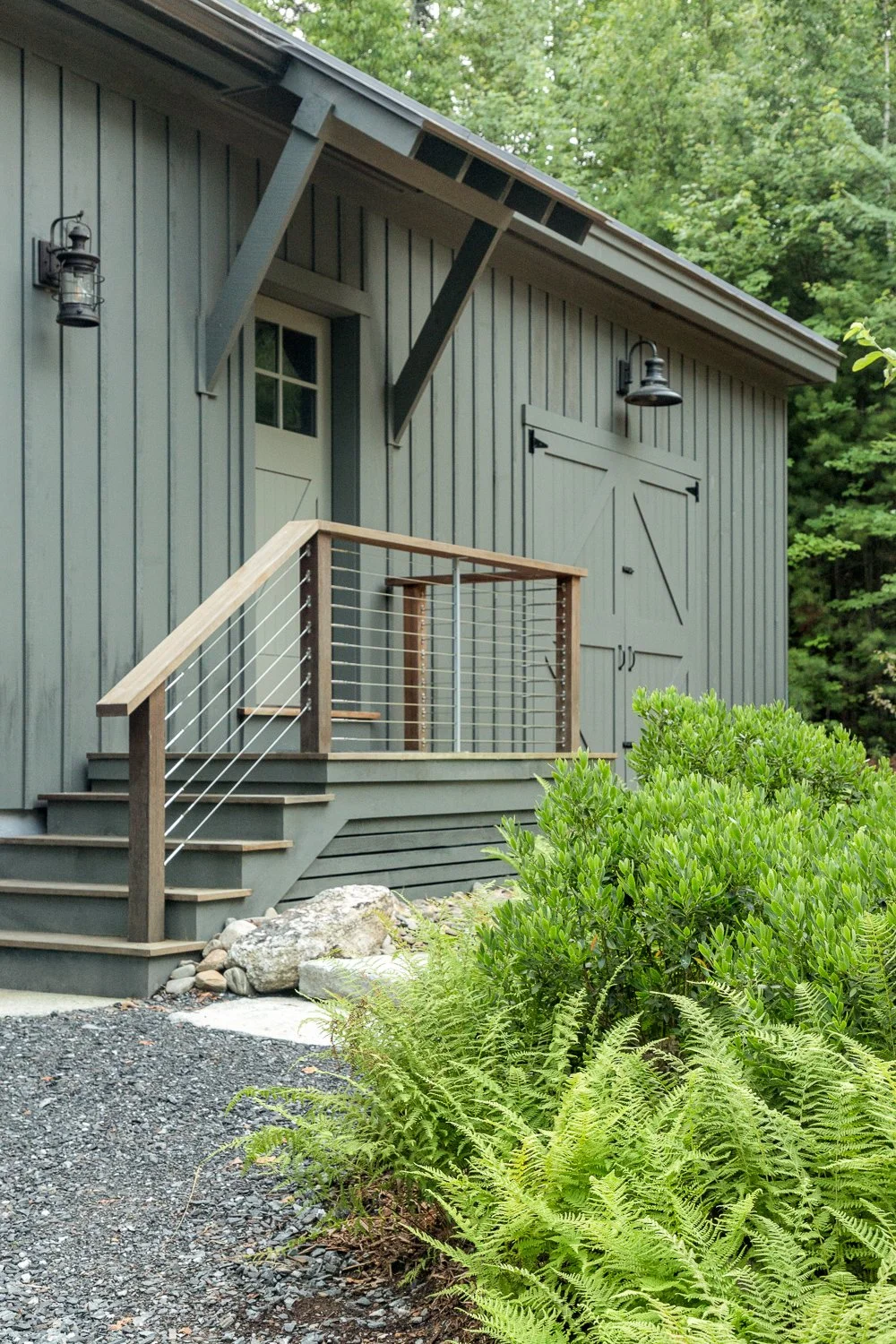 Gray building with a small deck and railing, surrounded by green bushes and ferns, with a gravel pathway in front.