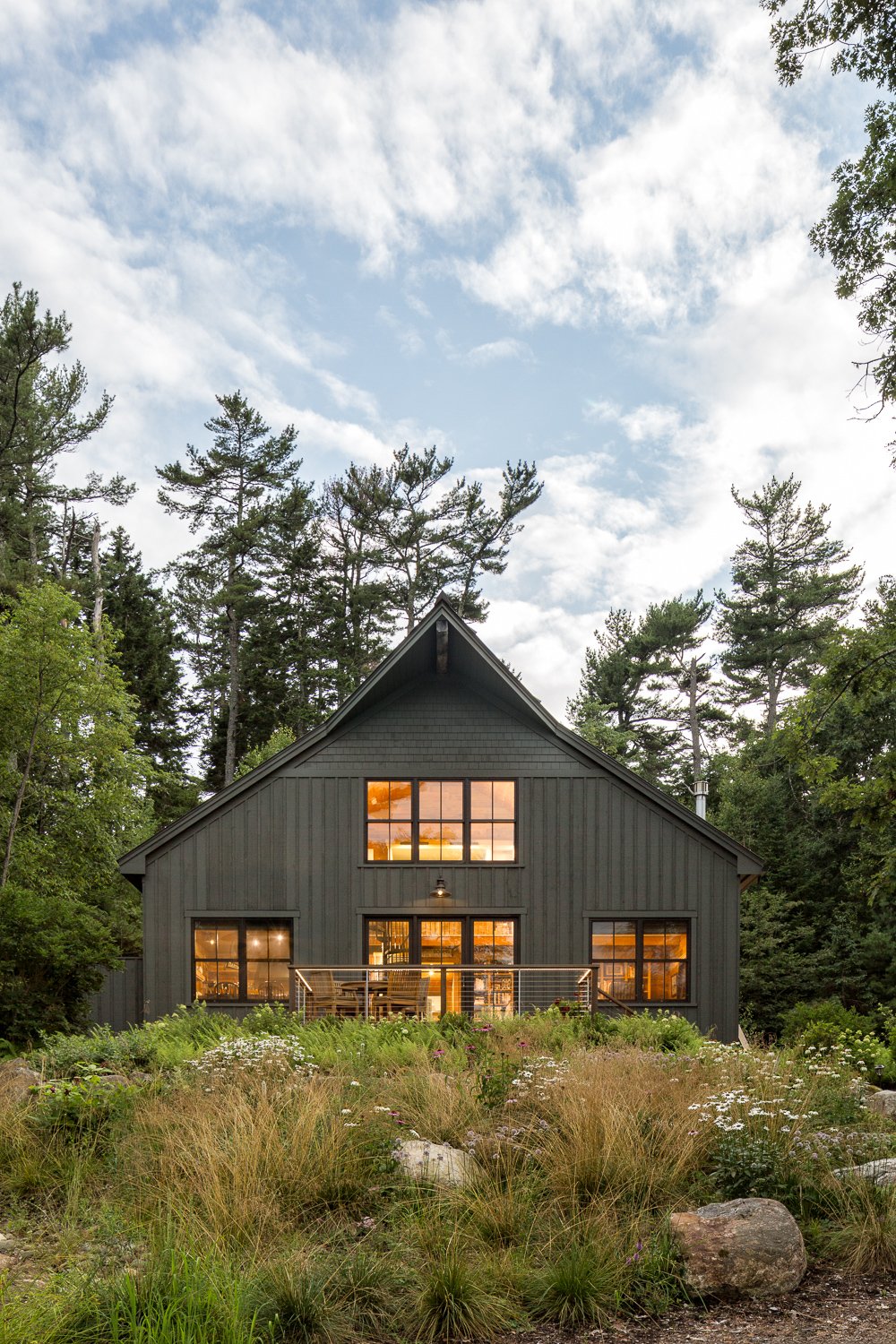 A modern dark gray house with large windows, set in a forested area with tall trees. The house has a balcony and is illuminated from inside, with a cloudy sky overhead.