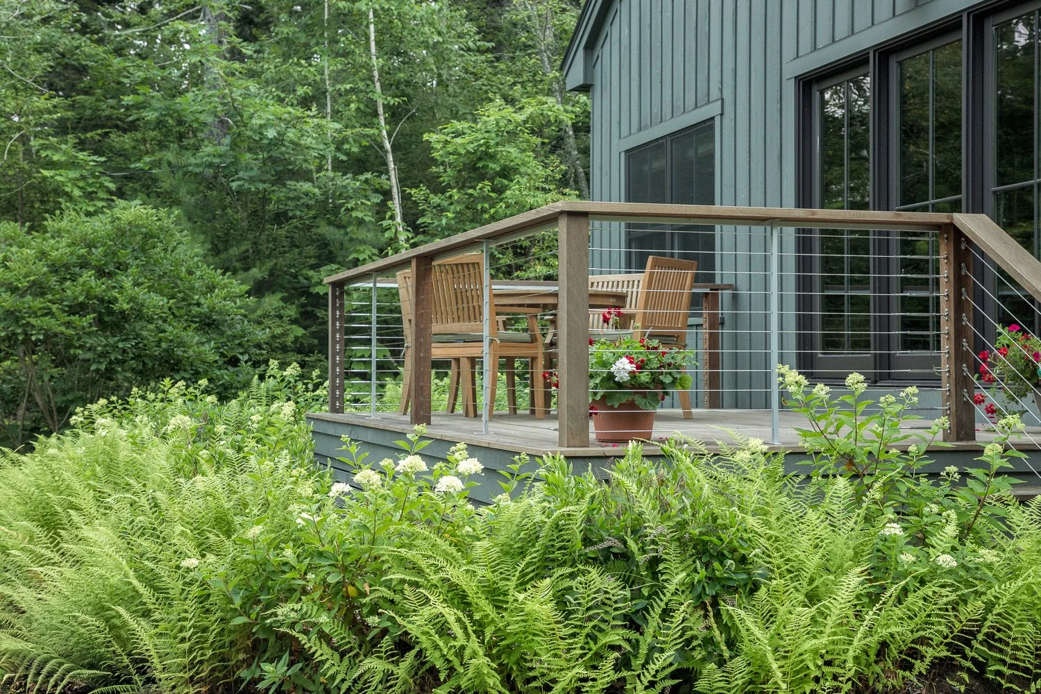 A wooden deck with outdoor furniture, potted flowers, and a railing, attached to a blue house in a lush green, wooded area.