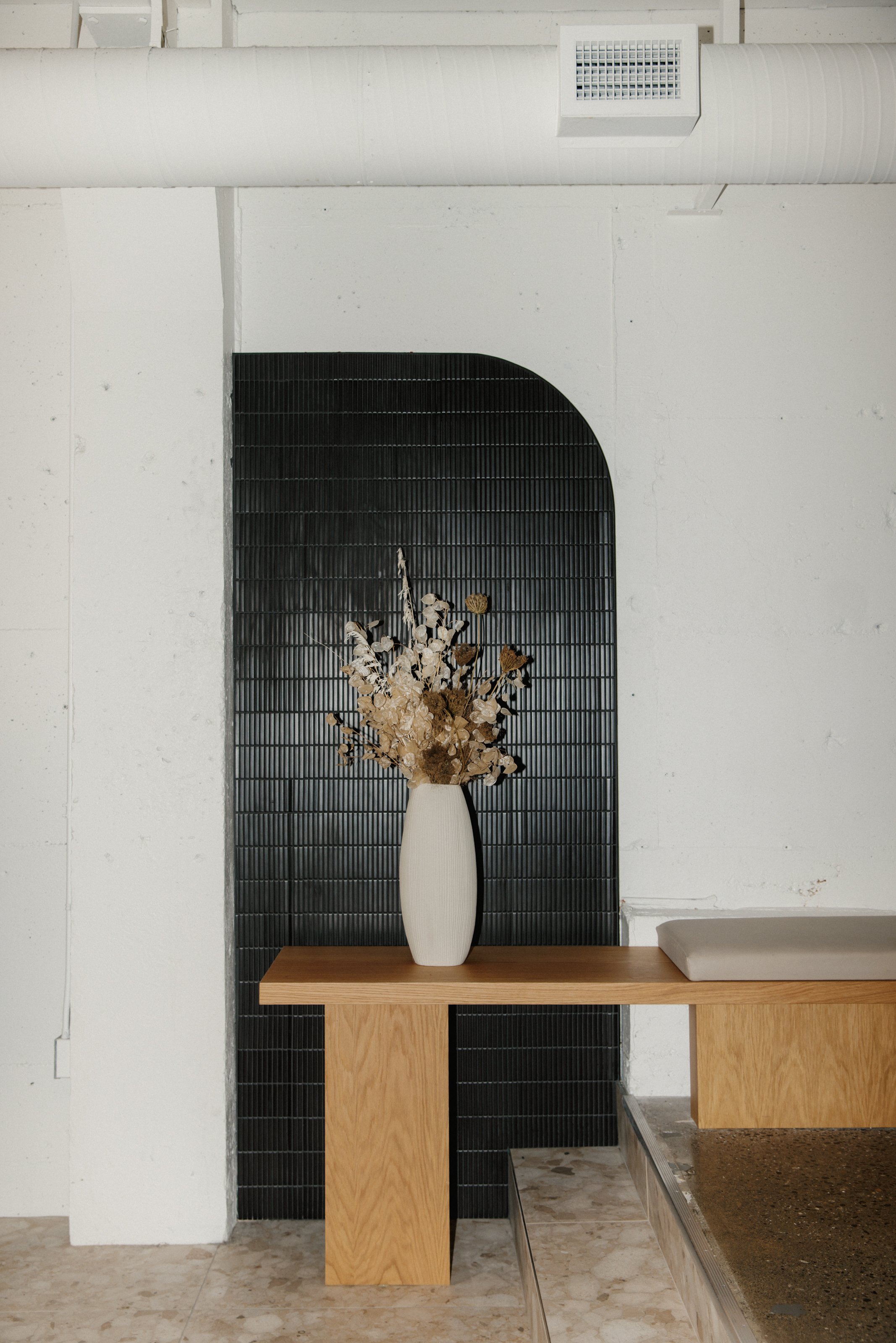 High flash image of millwork bench with vase of flowers and curved black tile behind.