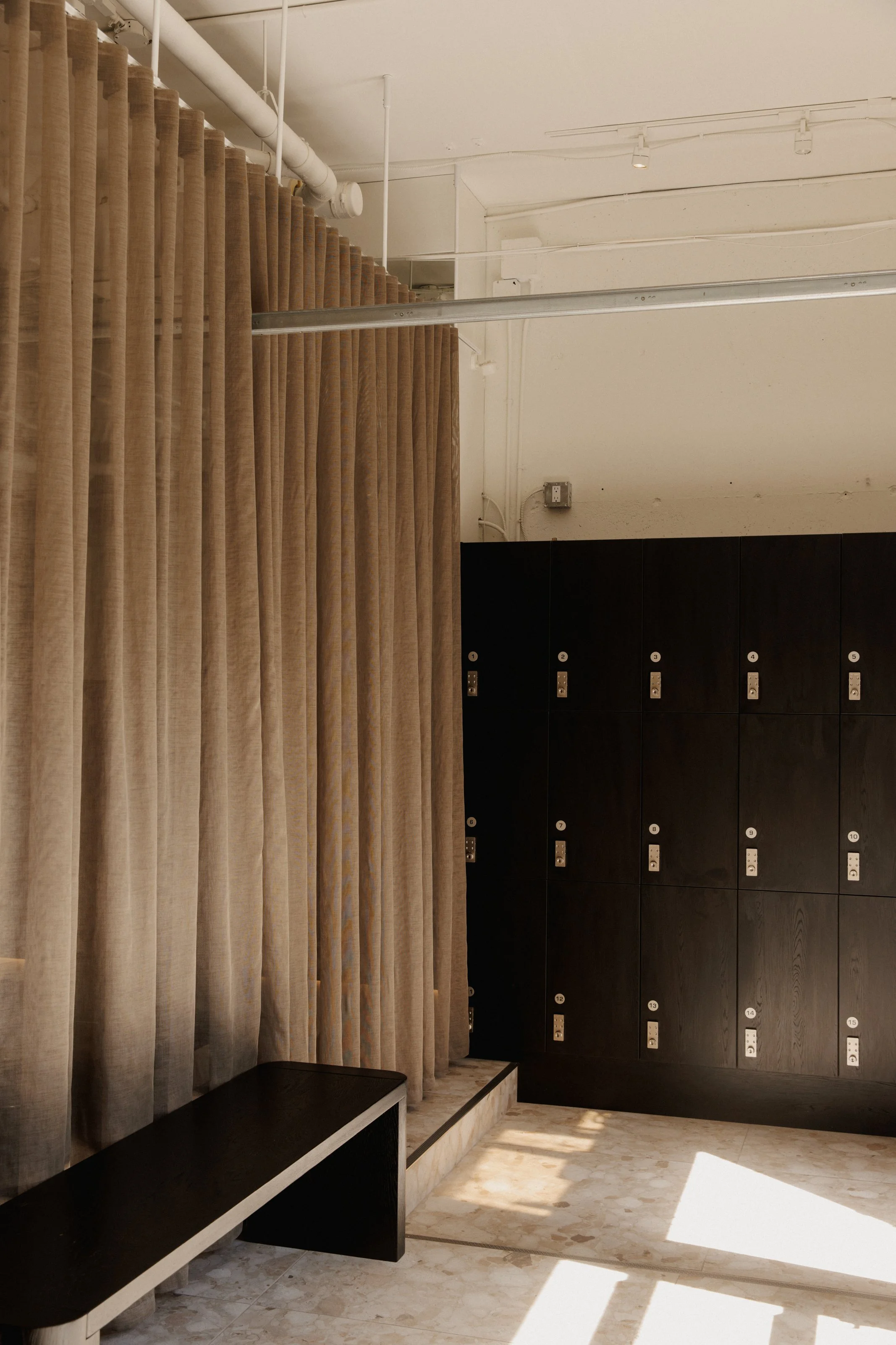 Entrance of studio with natural light shining in creating shadows on the beige tile floor. A sheer beige curtain separates the space. 15 black lockers in the background of the photo.